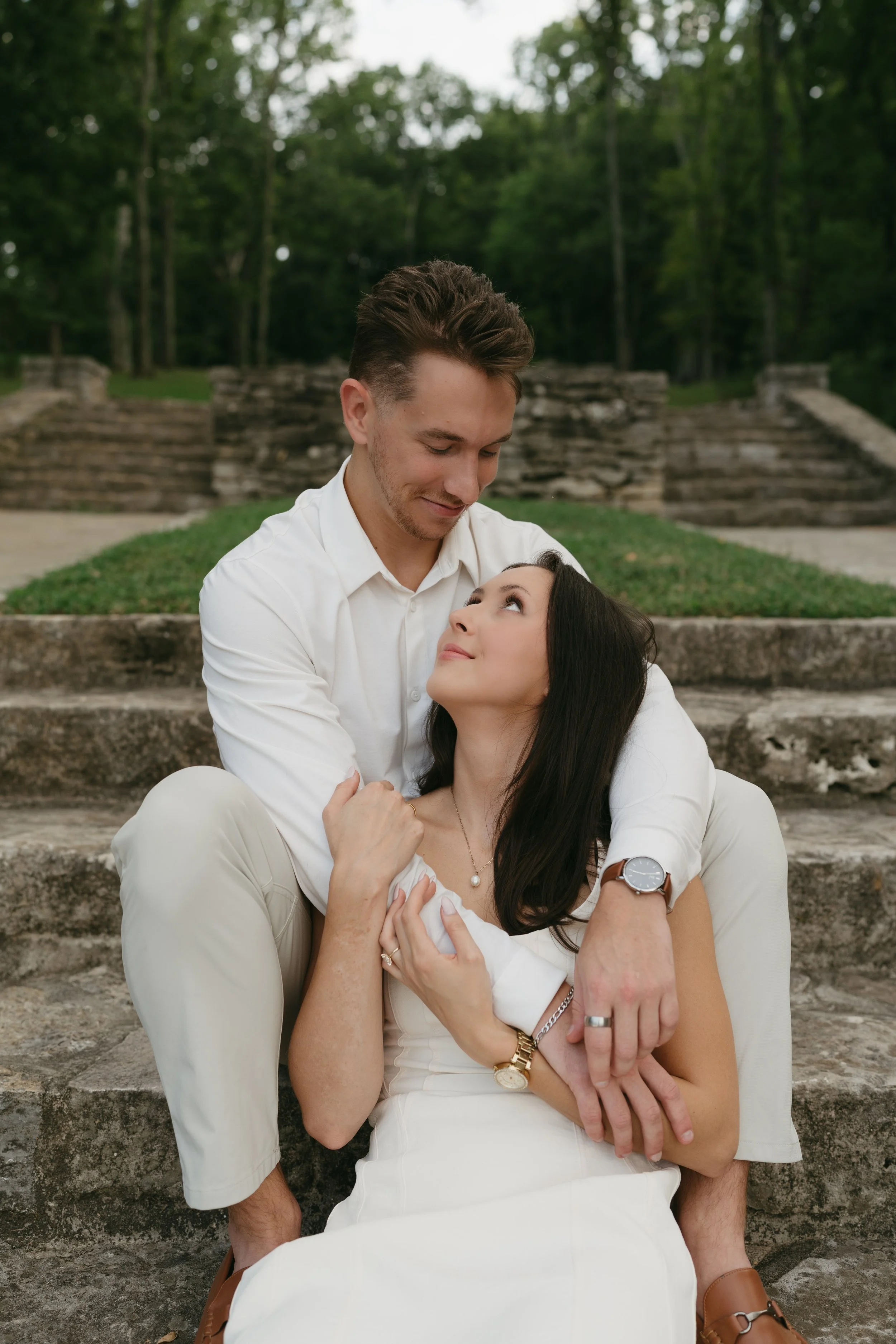 A man and a woman sitting on stone steps outdoors, gazing into each other's eyes, surrounded by greenery and trees in the background.