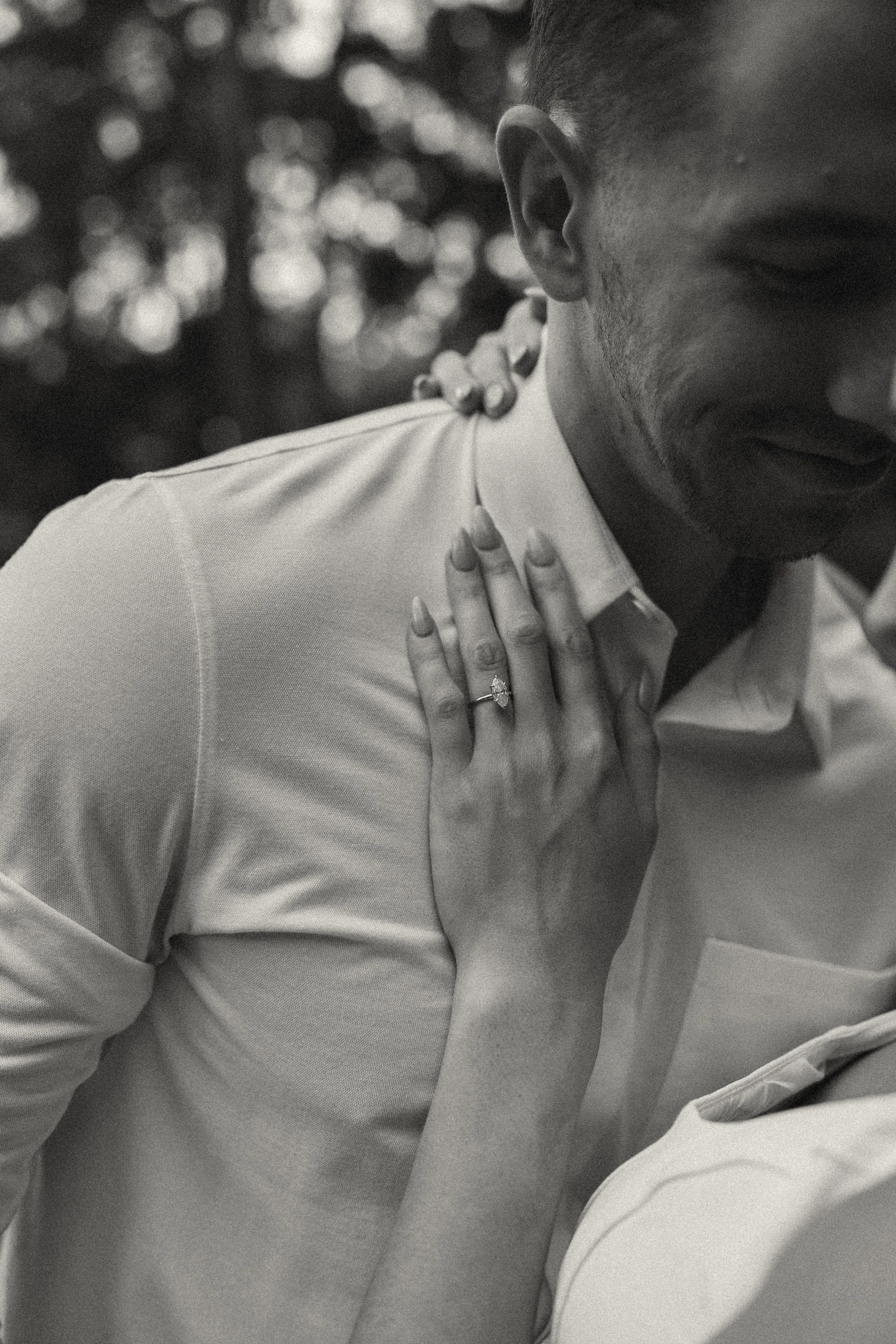 A close-up of a couple, man and woman, smiling and embracing, with the woman's hand showing an engagement ring on her finger, in black and white.