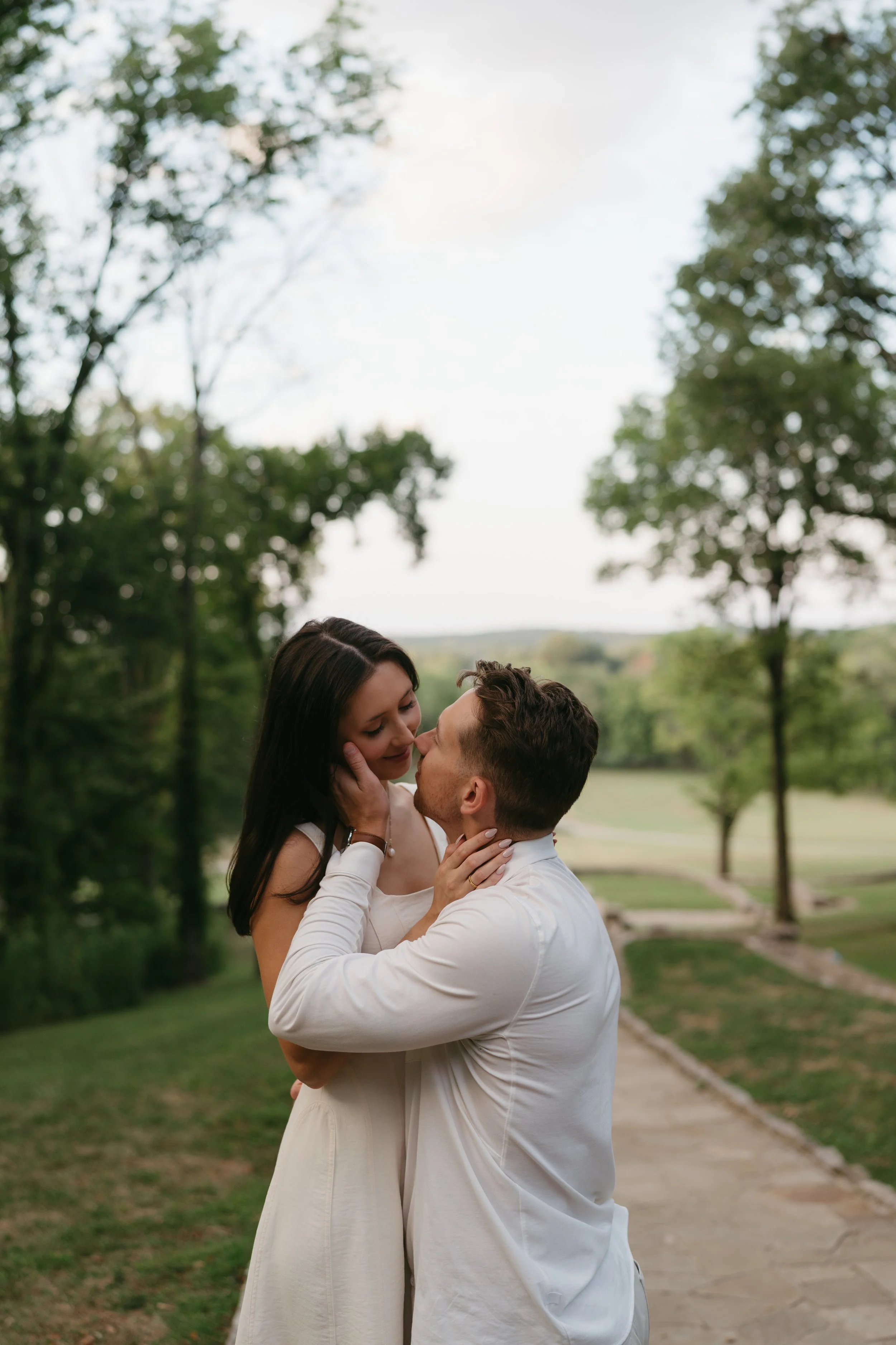 A couple kissing outdoors on a park path with green trees in the background.