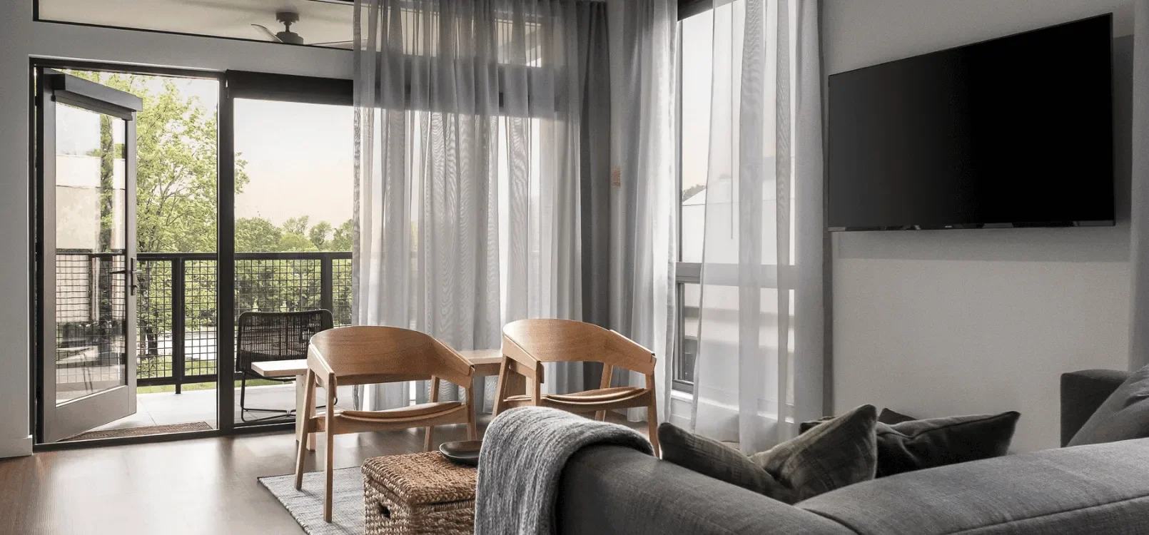Living room with a gray sofa, TV mounted on the wall, dining table with two wooden chairs, sheer white curtains, sliding glass door leading to a balcony with a view of trees.