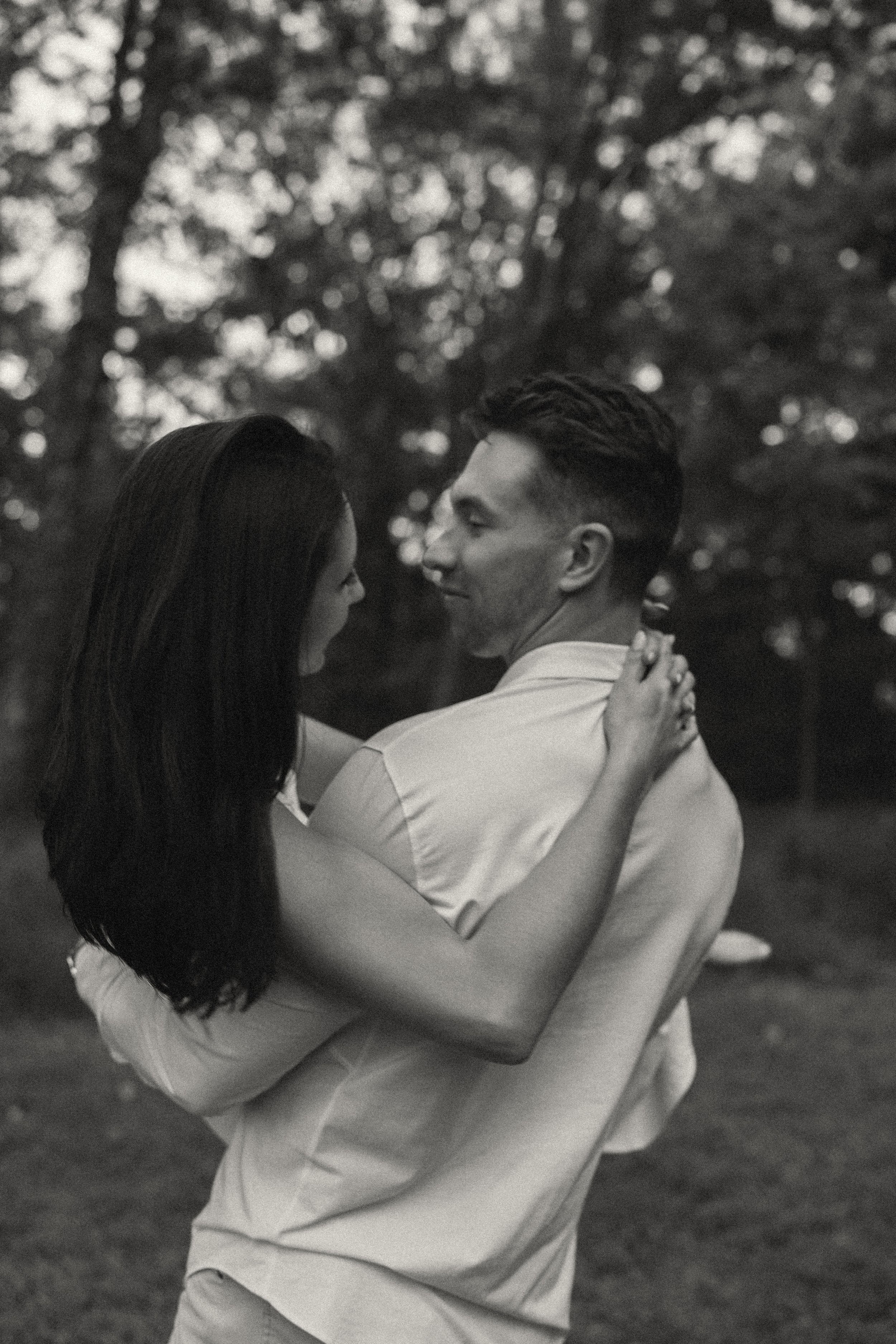 A black-and-white photo of a young couple dancing outdoors, smiling and gazing at each other, with trees blurred in the background.