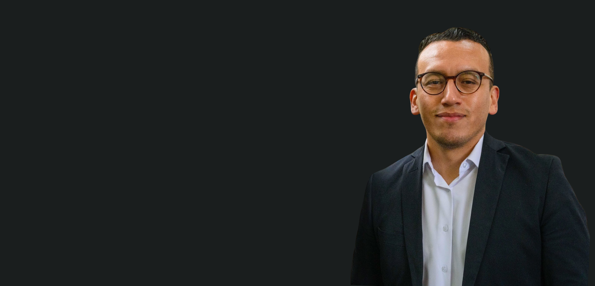 A portrait of a young man with glasses, wearing a dark suit and white shirt, standing against a dark background.