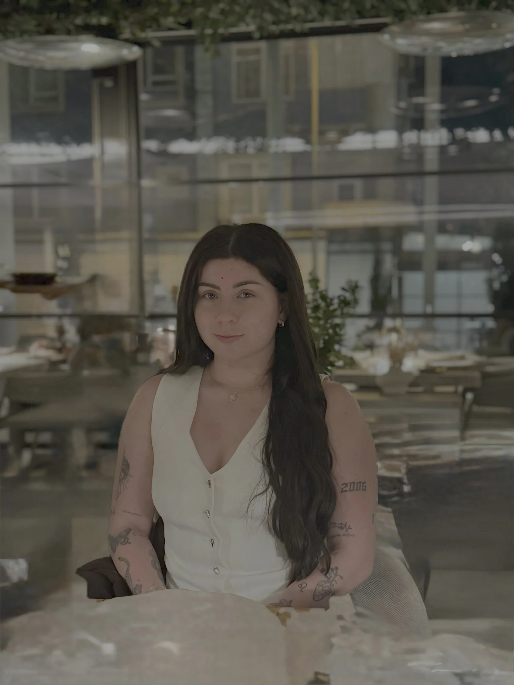 A woman with long dark hair and tattoos on her arms, sitting at a table in a modern restaurant or cafe, with large windows in the background revealing reflections of the interior and street outside.