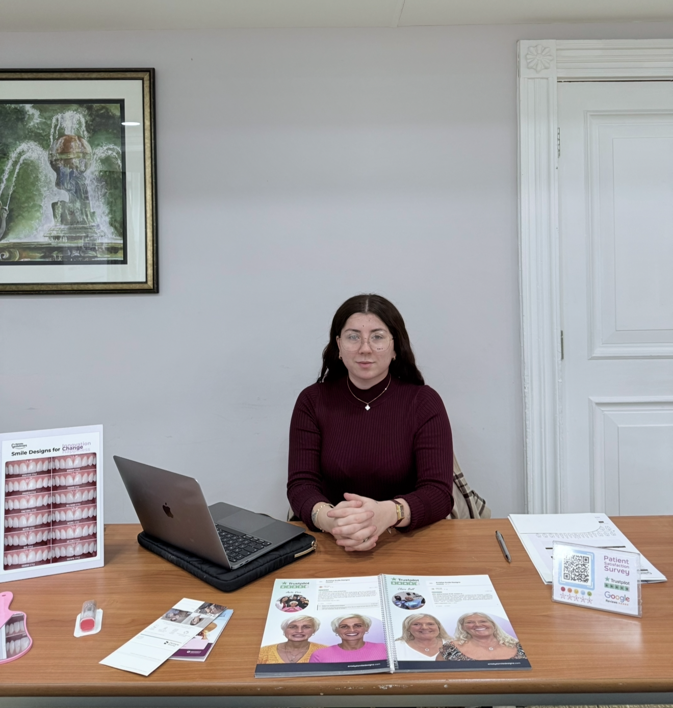 A woman sitting at a wooden desk with dental brochures, a laptop, and a patient survey sign displayed.