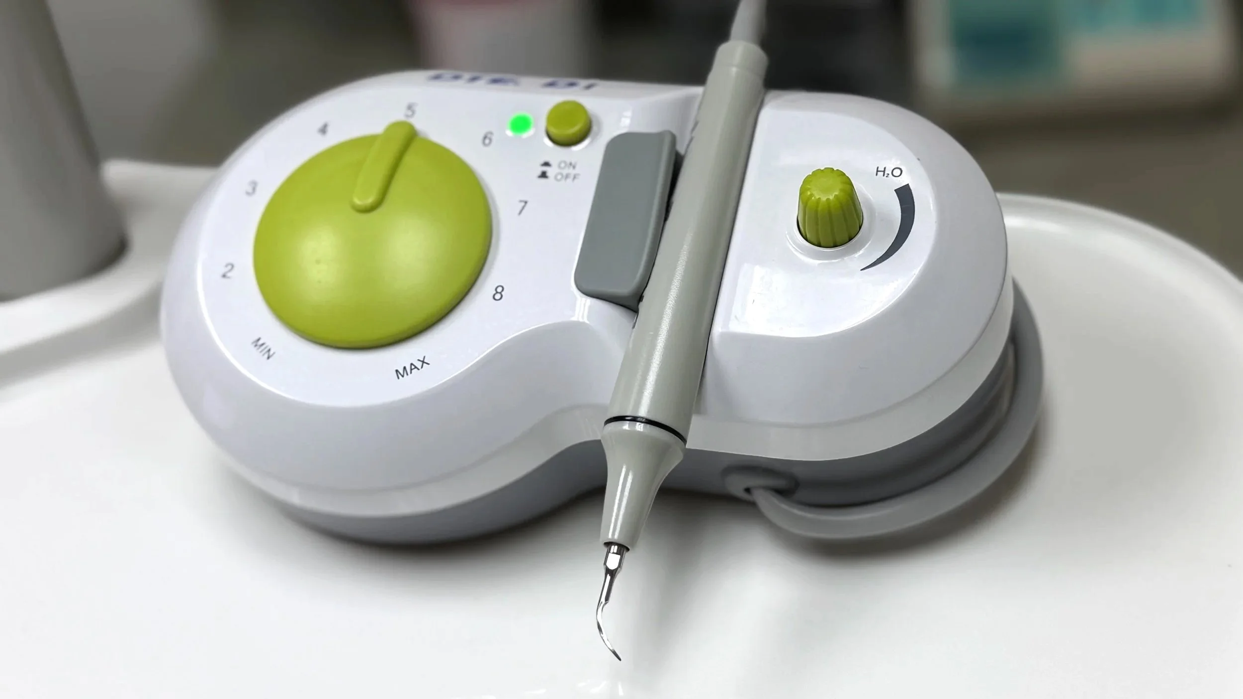 Periodontal ultrasonic scaler on a white machine with a gray handpiece, a green control knob, and a small button, used for scaling procedures