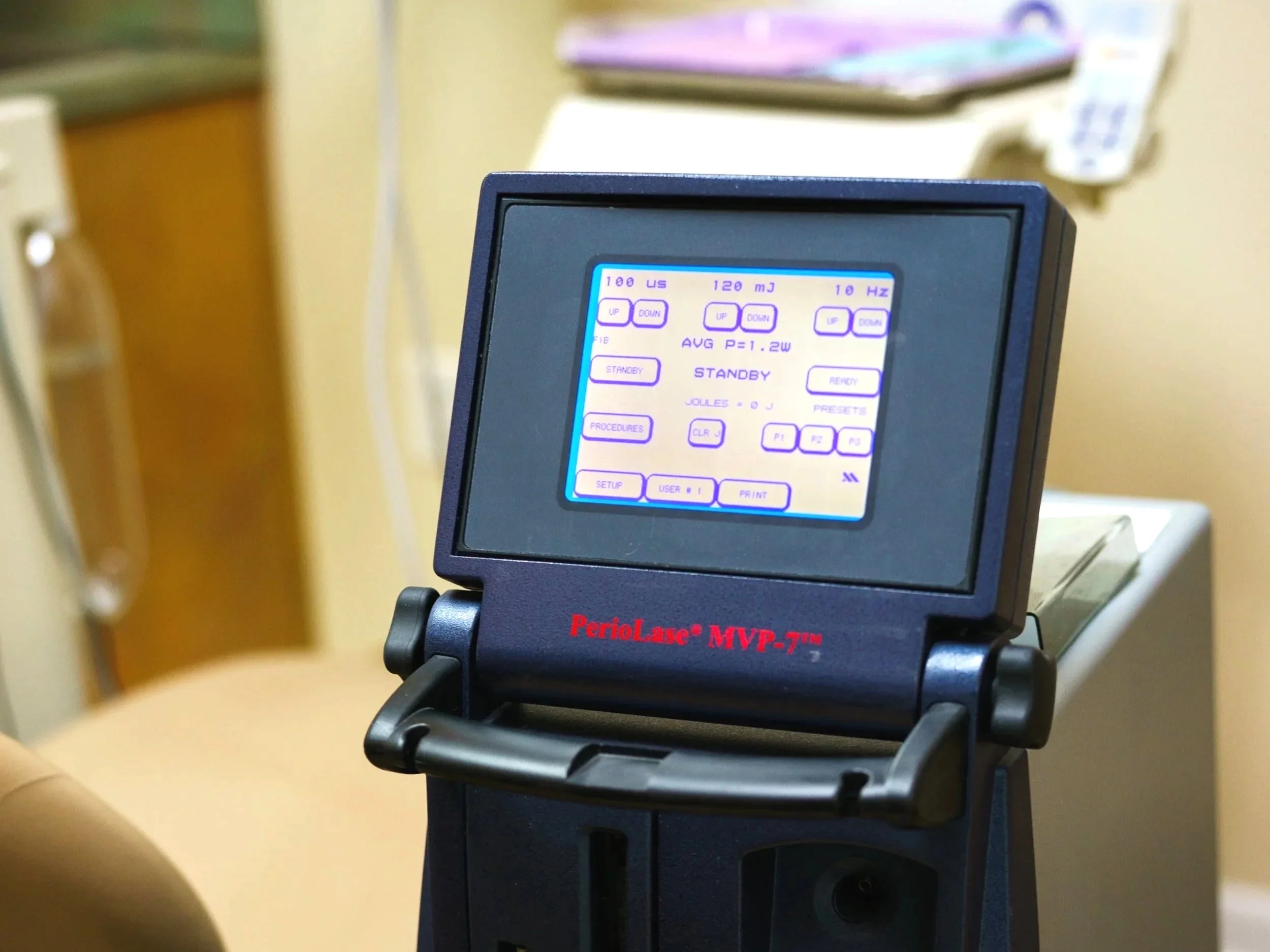 Medical monitor screen displaying settings for a dental laser, labeled 'PerioLase MVP-7', with buttons for adjusting parameters, and a background with a yellow wall, and a dental chair and instruments in the background.
