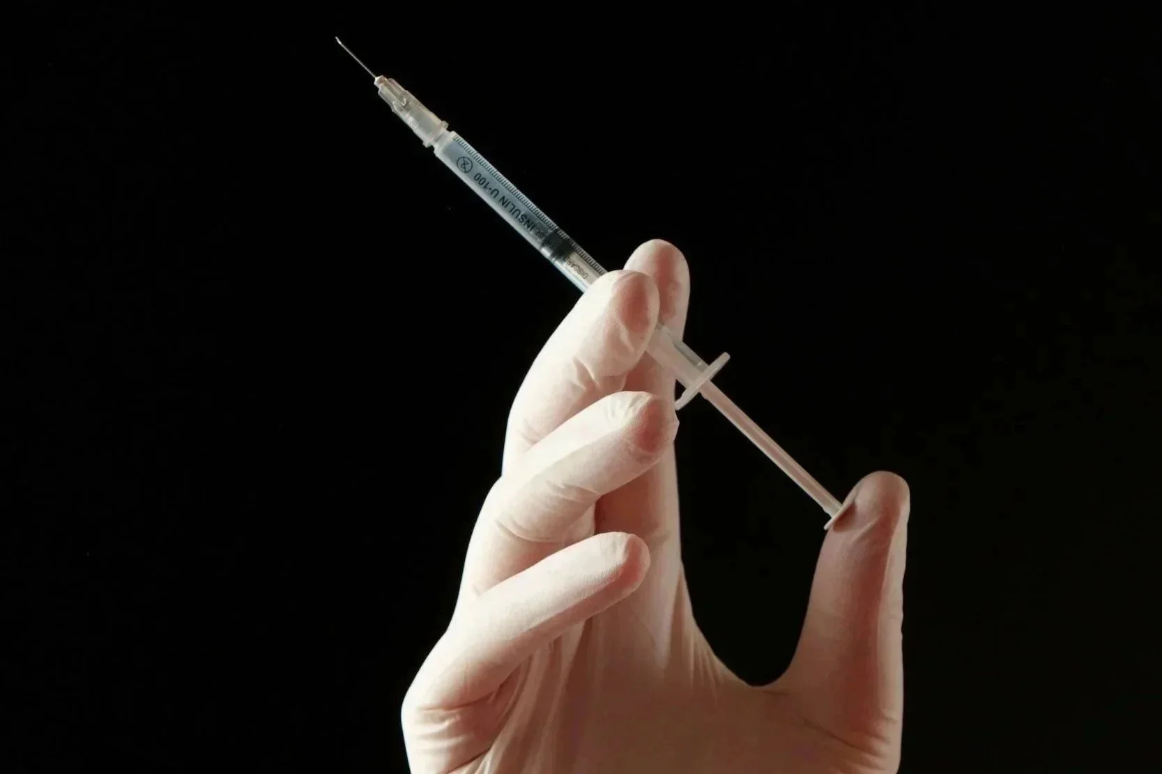 A gloved hand holding a syringe against a black background.