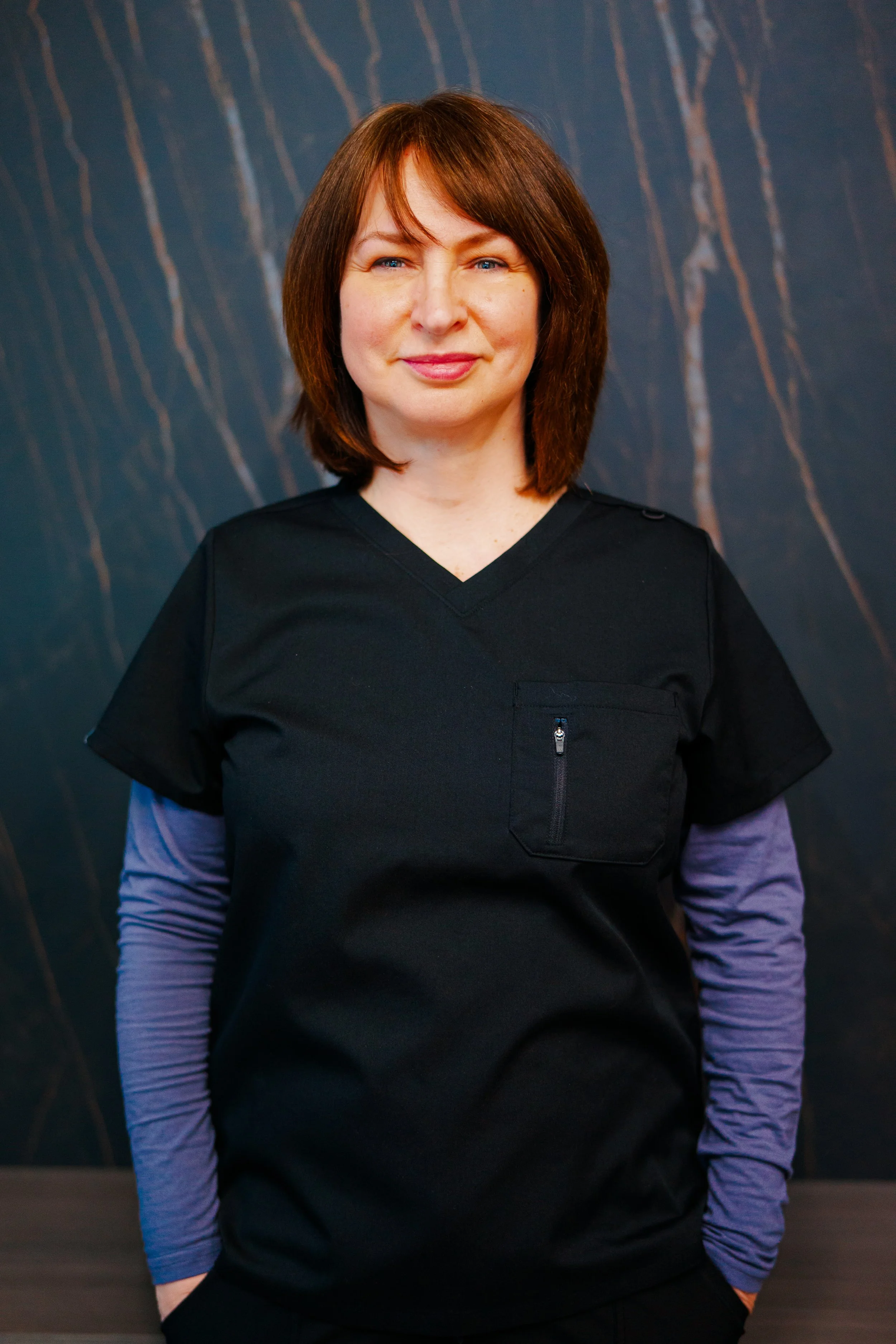 A woman with shoulder-length brown hair, wearing black medical scrubs with a blue long-sleeve undershirt, standing against a dark marble background.
