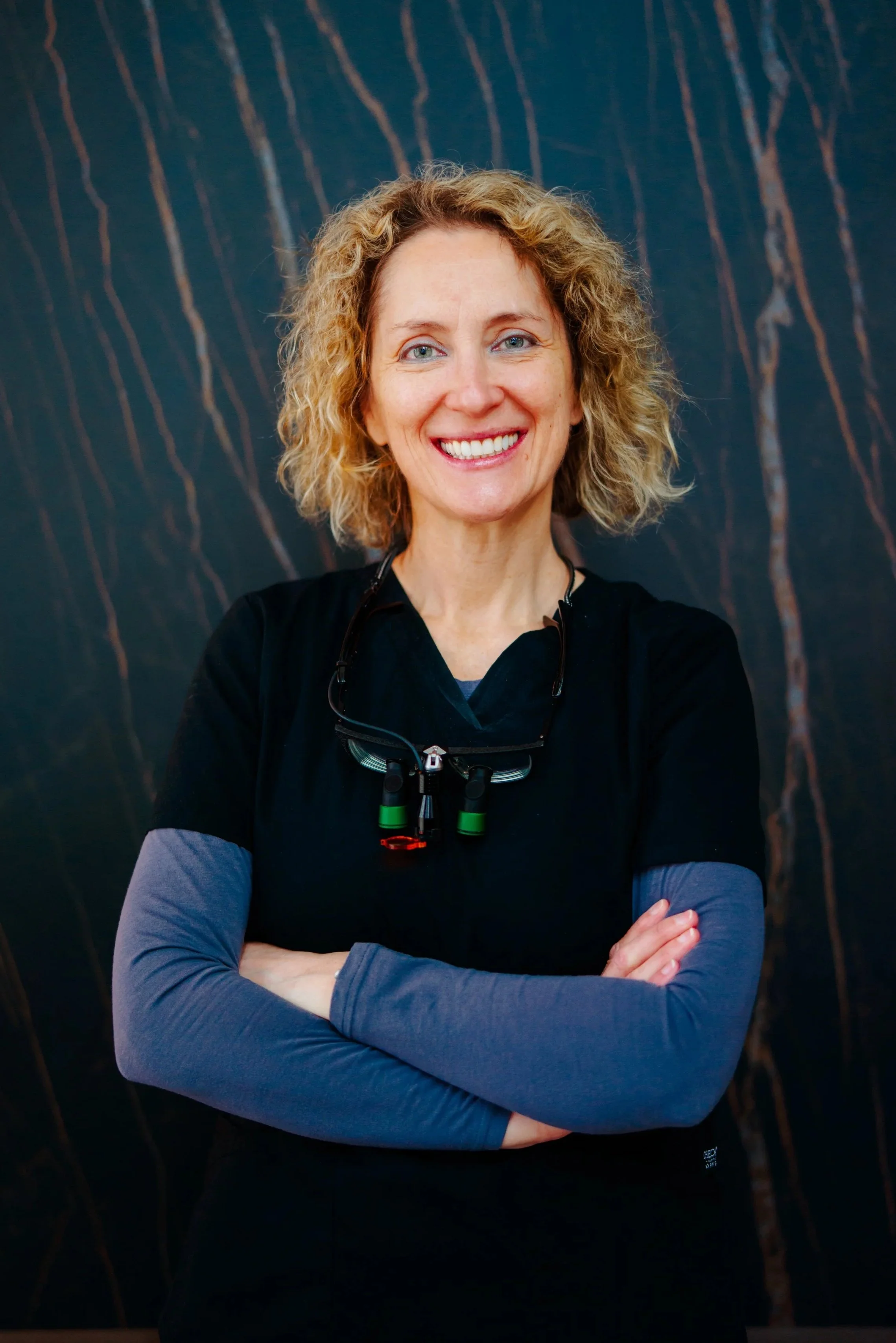 A smiling woman with curly blonde hair wearing black scrubs with a gray long sleeve underneath. She has loops standing in front of a dark marbled background.