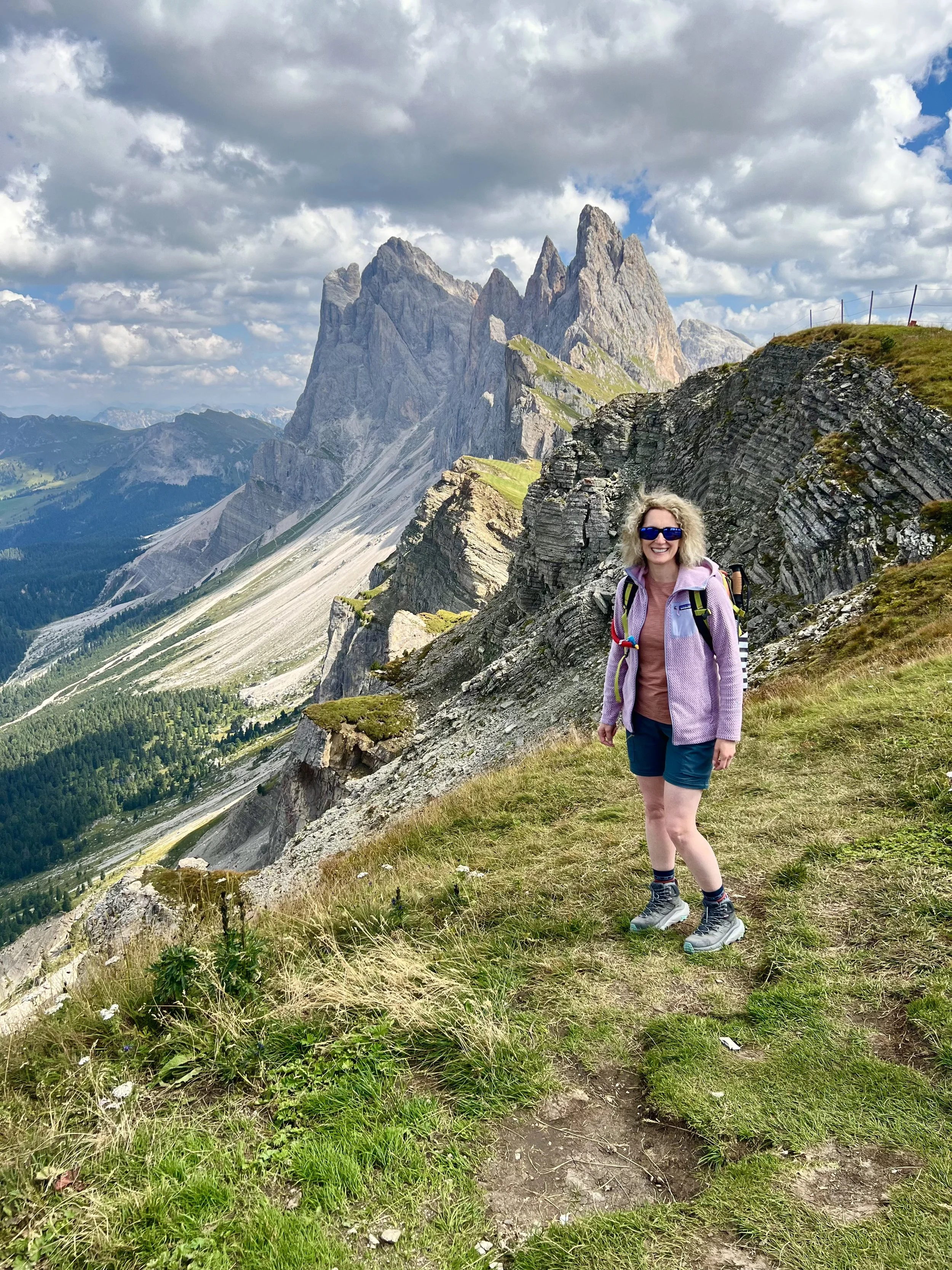 Woman hiking in the mountains wearing sunglasses, a purple jacket, and shorts, with a backpack. The background features rugged mountain peaks, grassy slopes, and a partly cloudy sky.