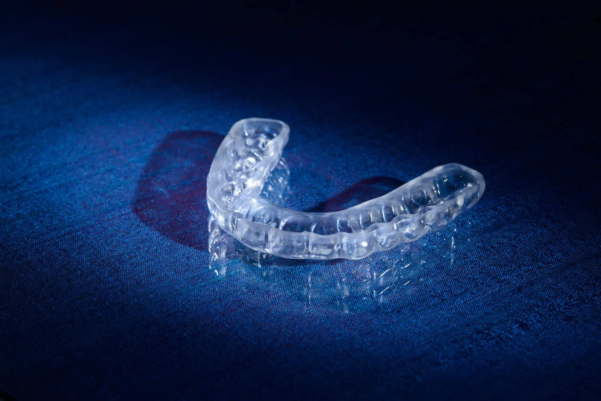 A clear dental night guard resting on a blue fabric background, casting a shadow.