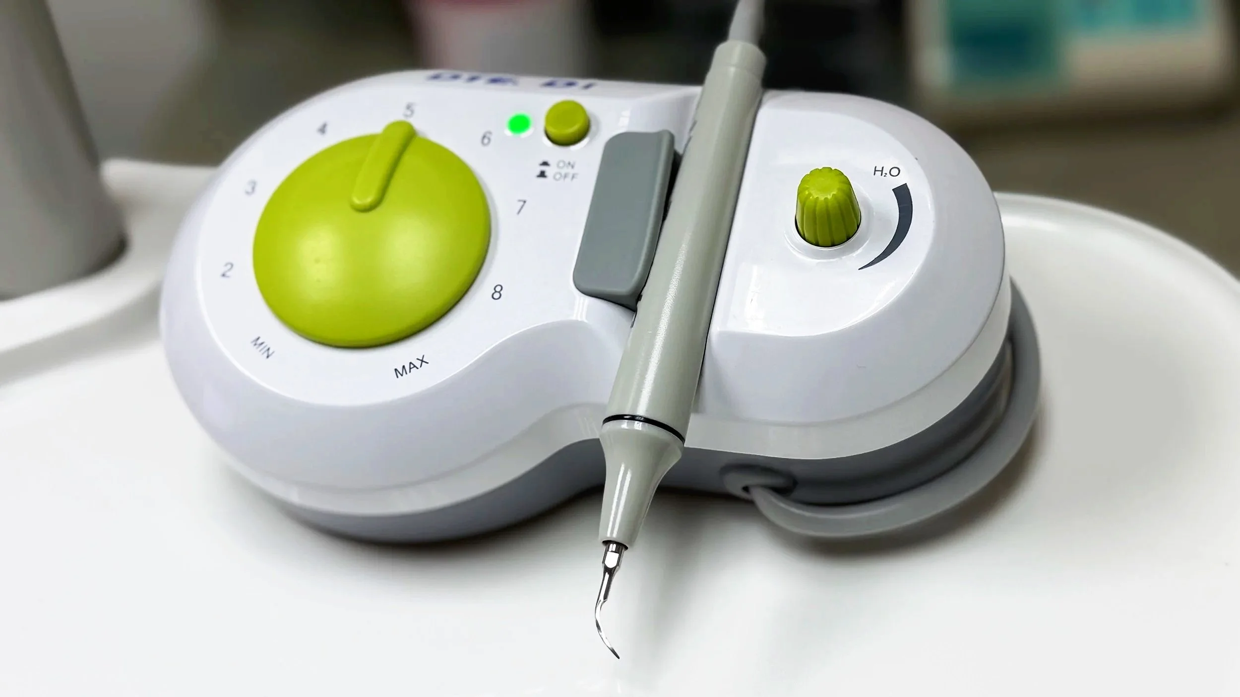 A dental ultrasonic scaler with a green dial, a grey handle, and a water spray control on a dental tray.