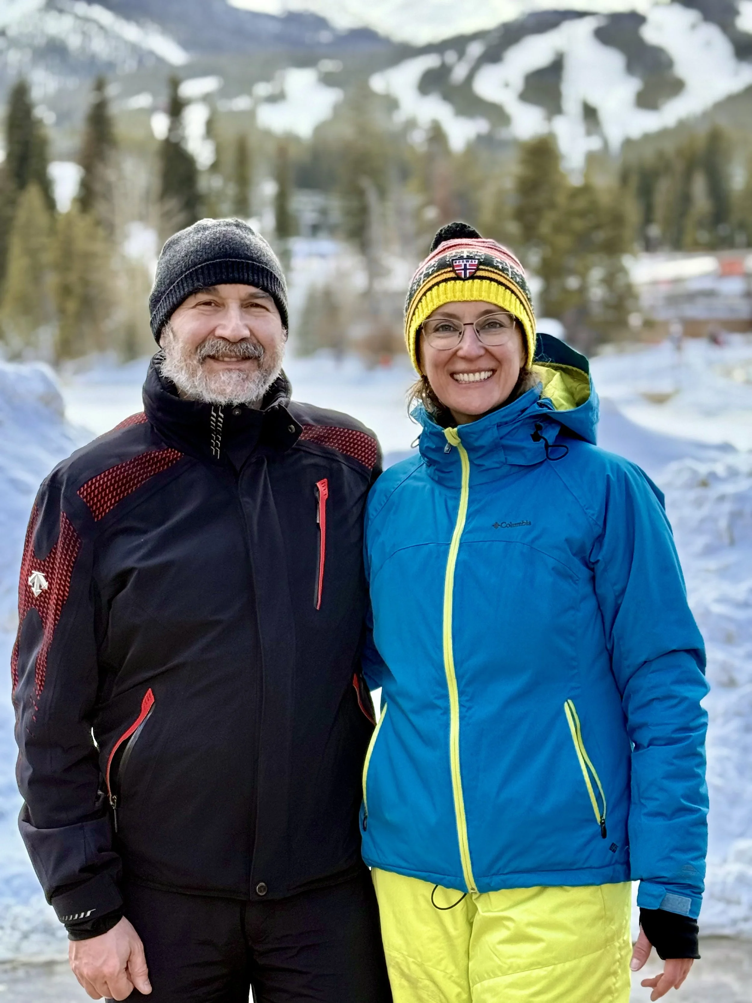 A smiling man and woman dressed in ski outfits and knit hats standing outdoors in a snowy mountain landscape.