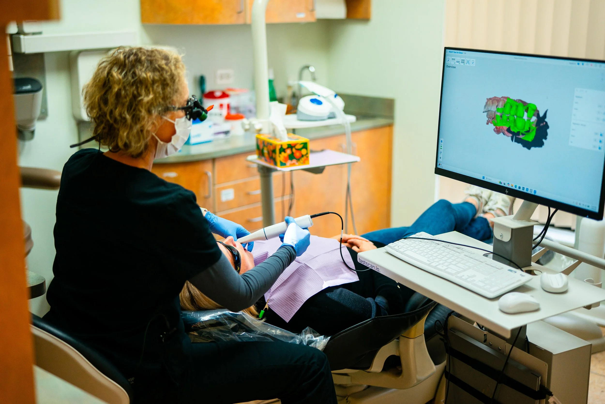 Dental professional using a 3D scanner on a patient's mouth in a dental clinic, with a computer screen displaying a live-scanned 3D model of teeth.