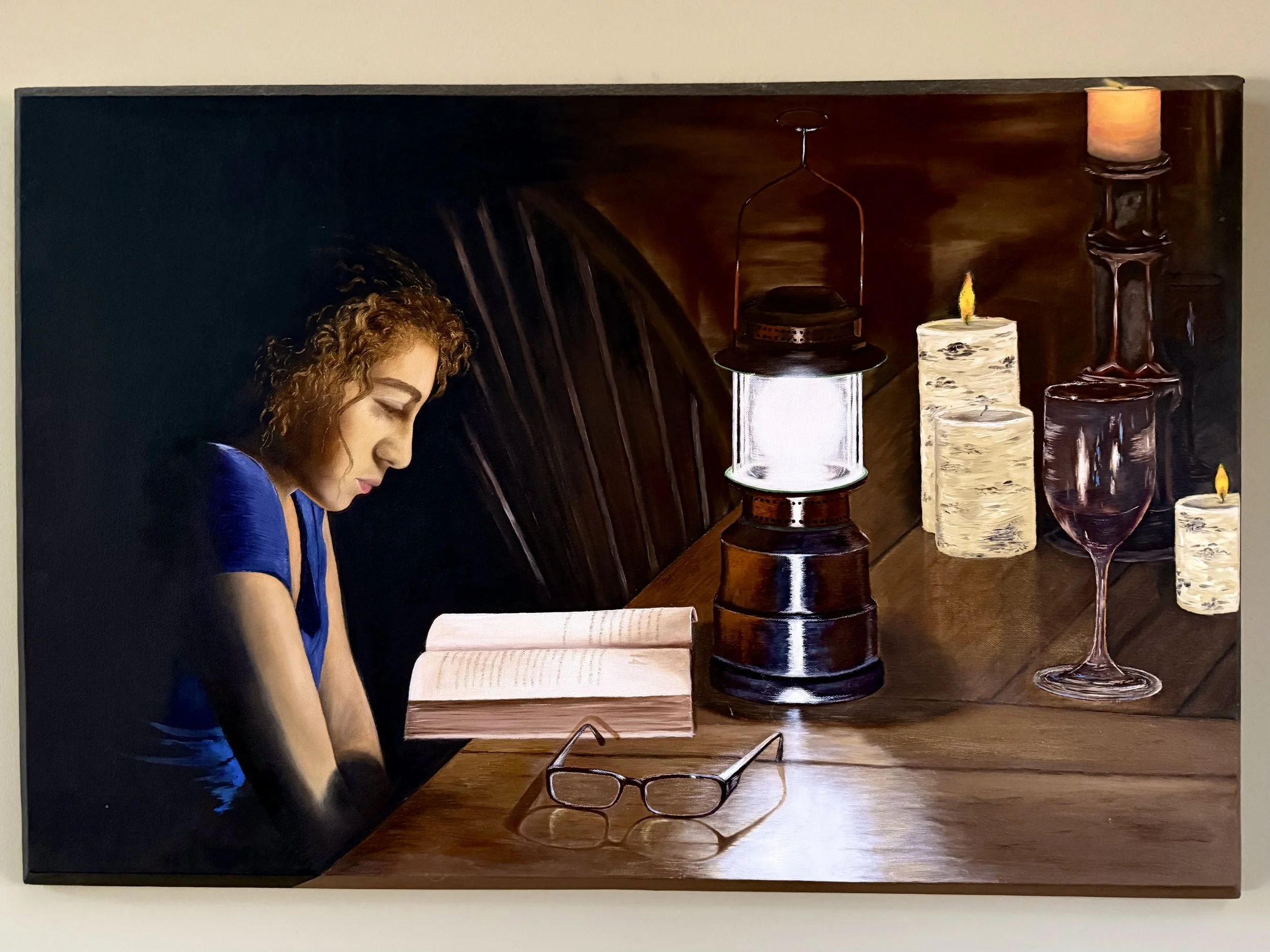Realistic painting of a teenage girl with curly hair wearing a blue top, reading a book at a wooden table with candles, a lantern, glasses, and wine glasses.