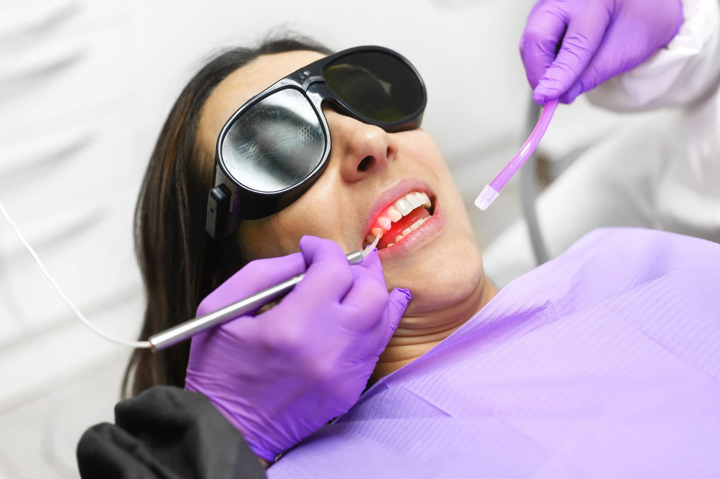A woman with safety glasses and purple gloves lying in a dental chair, undergoing a dental procedure involving a dental scaler and suction device.