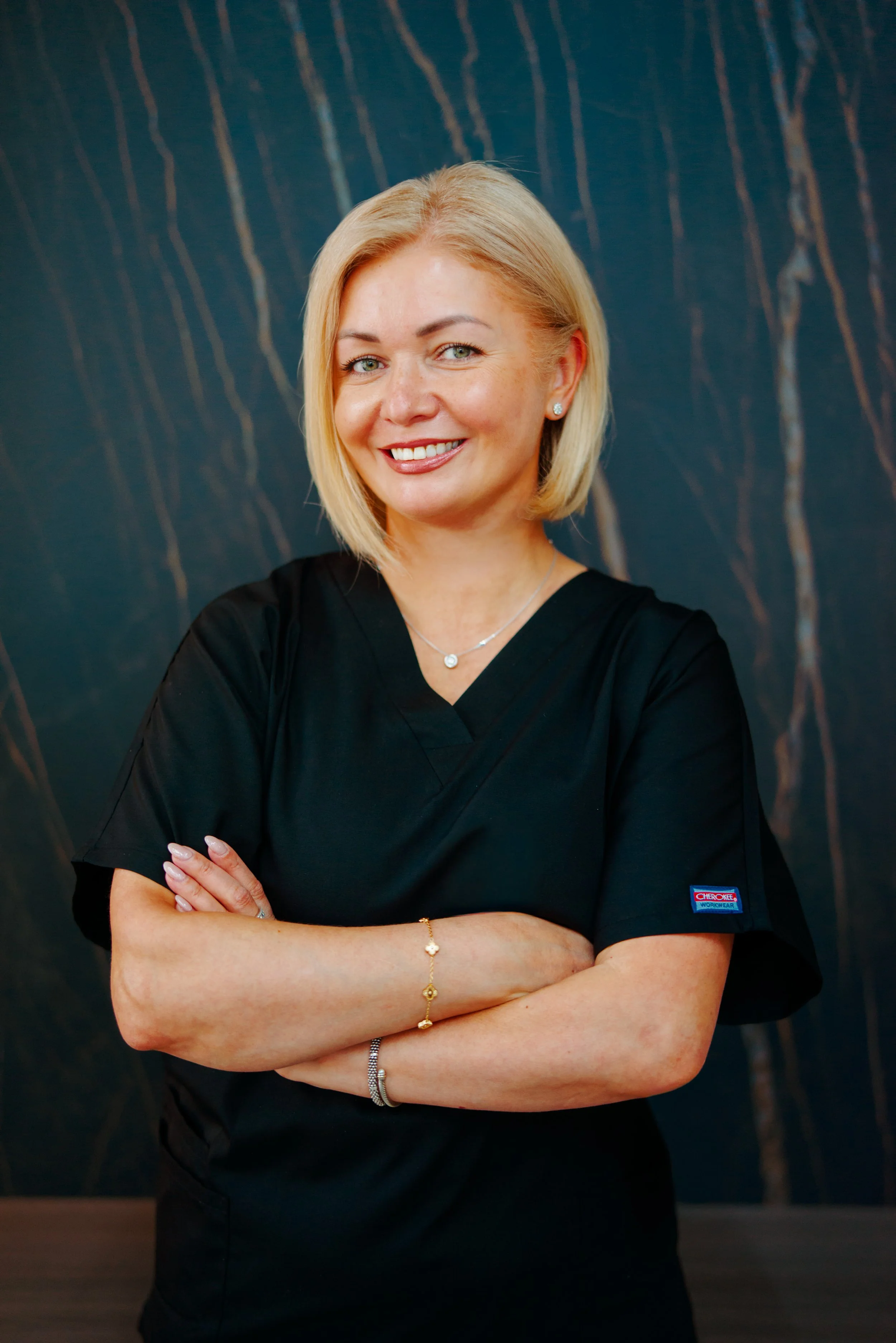 A woman with blonde hair, wearing black scrubs, standing with arms crossed and smiling, against a dark background with streaks of brown and gold.