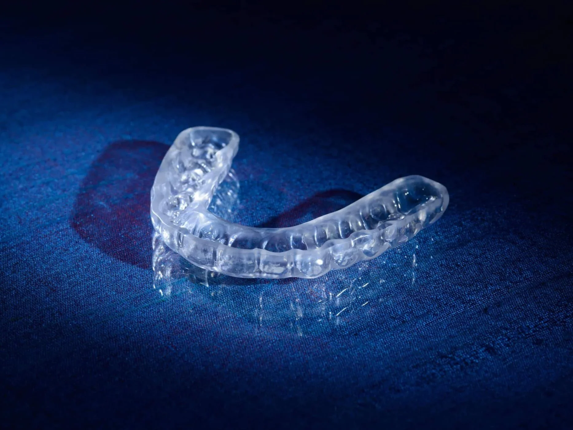 A clear dental night guard on a dark blue surface with lighting creating reflections.