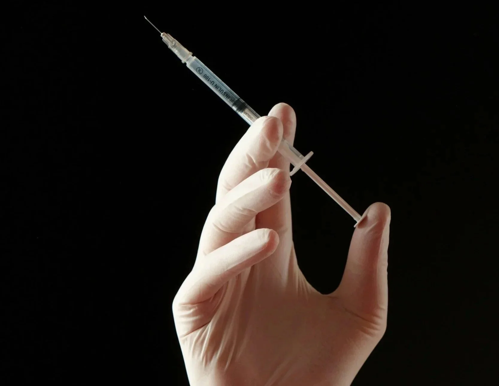 A gloved hand holding a syringe filled with a clear liquid, with a black background.