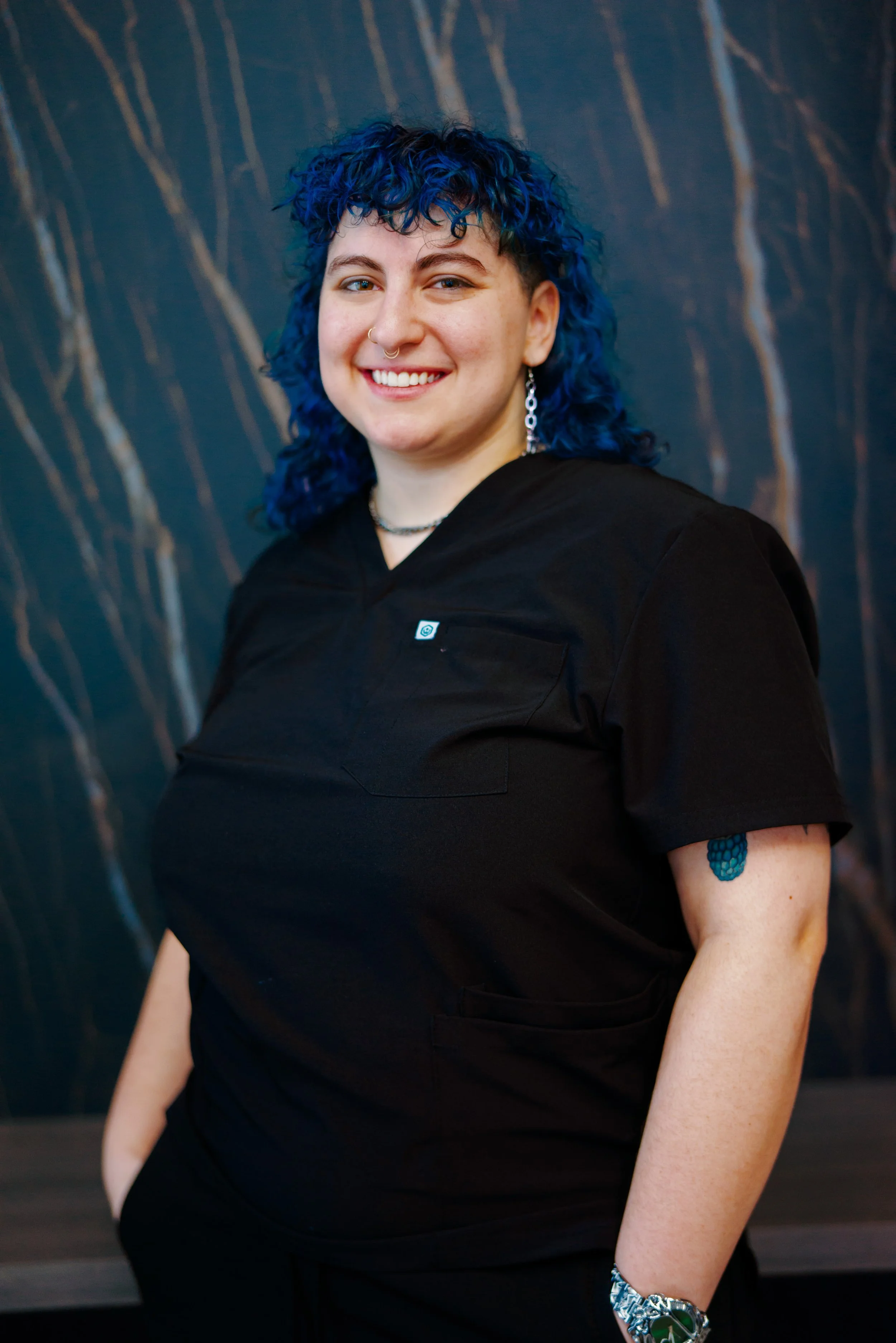 A smiling person with a blue, curly mullet and shaved sides. They are wearing black scrubs, standing in front of a dark marble wall. A tattoo of a blue raspberry peaks out under the short sleeve of the scrub.