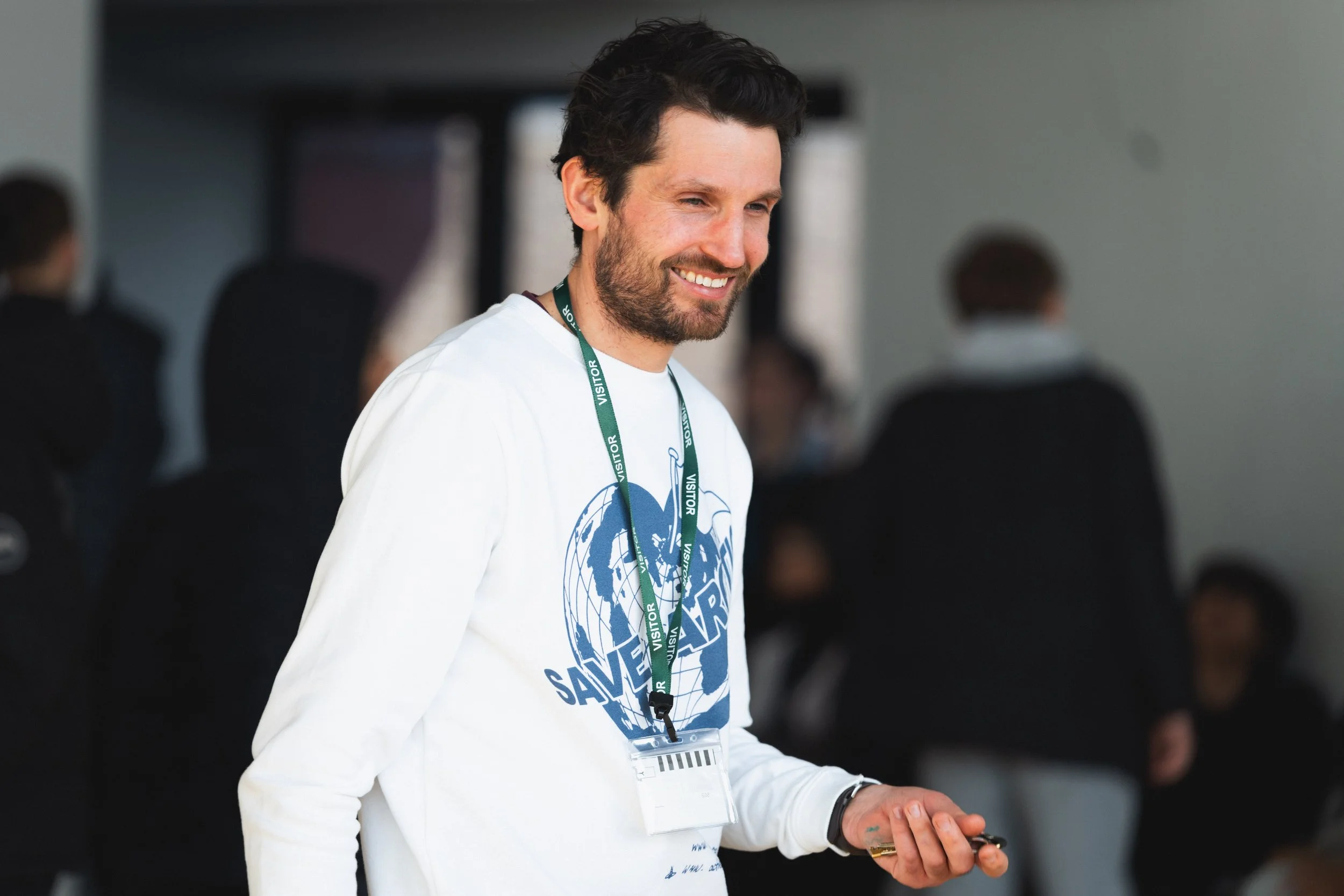 A smiling man with dark hair and a beard, wearing a white sweatshirt with a blue globe and shark graphic and a green visitor lanyard, holding a small object, in an indoor setting with blurred people in the background.