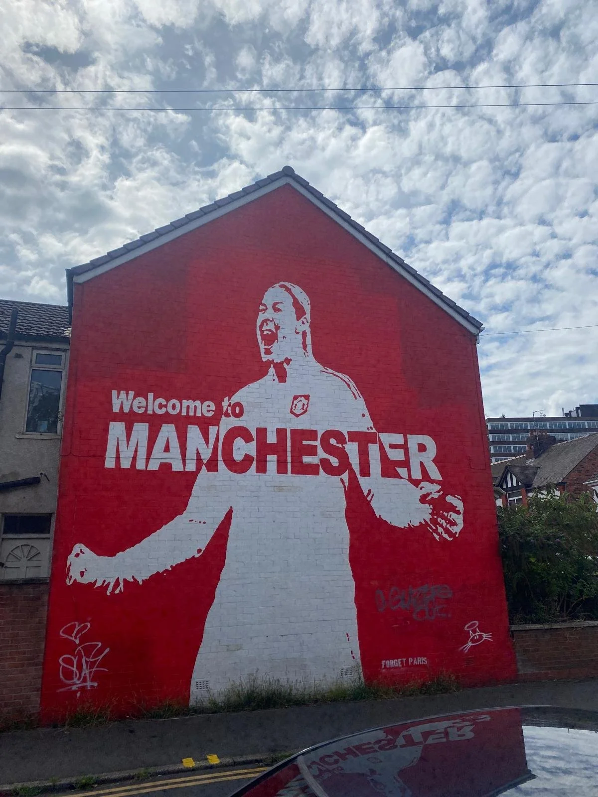 Street mural playing on a red wall with the words "Welcome to MANCHESTER" and an image of a person with an open mouth, arms outstretched, and a short hairstyle. The mural includes graffiti and tags, with a background of a partly cloudy sky.