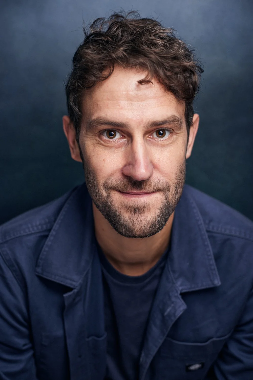 Close-up photo of a man with short, dark, curly hair and a beard, wearing a dark blue shirt, smiling slightly against a dark background.