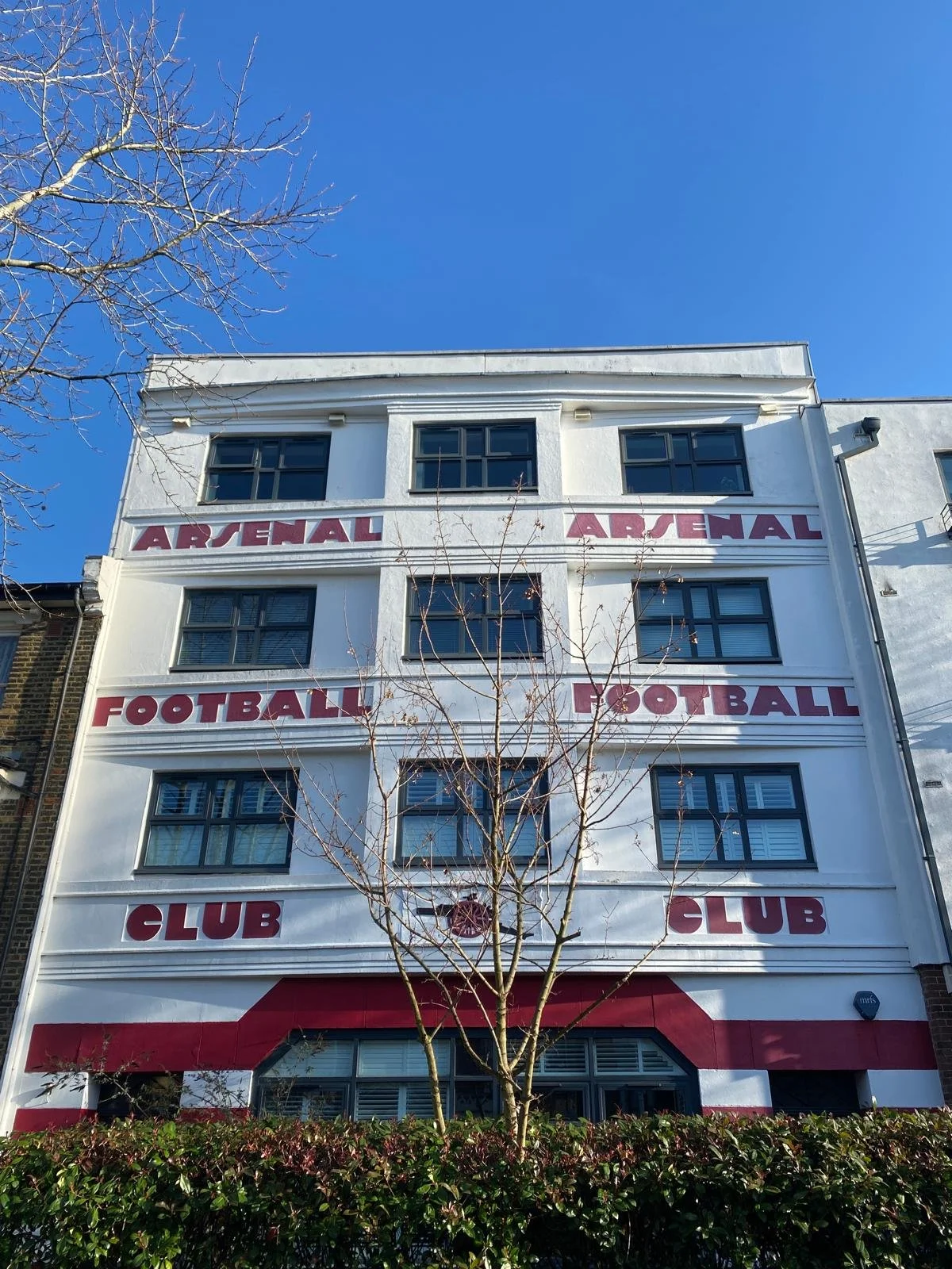 A white multi-story building with the words 'ARSENAL,' 'FOOTBALL,' and 'CLUB' painted in large red letters on the facade, with a small tree in front and a clear blue sky in the background.