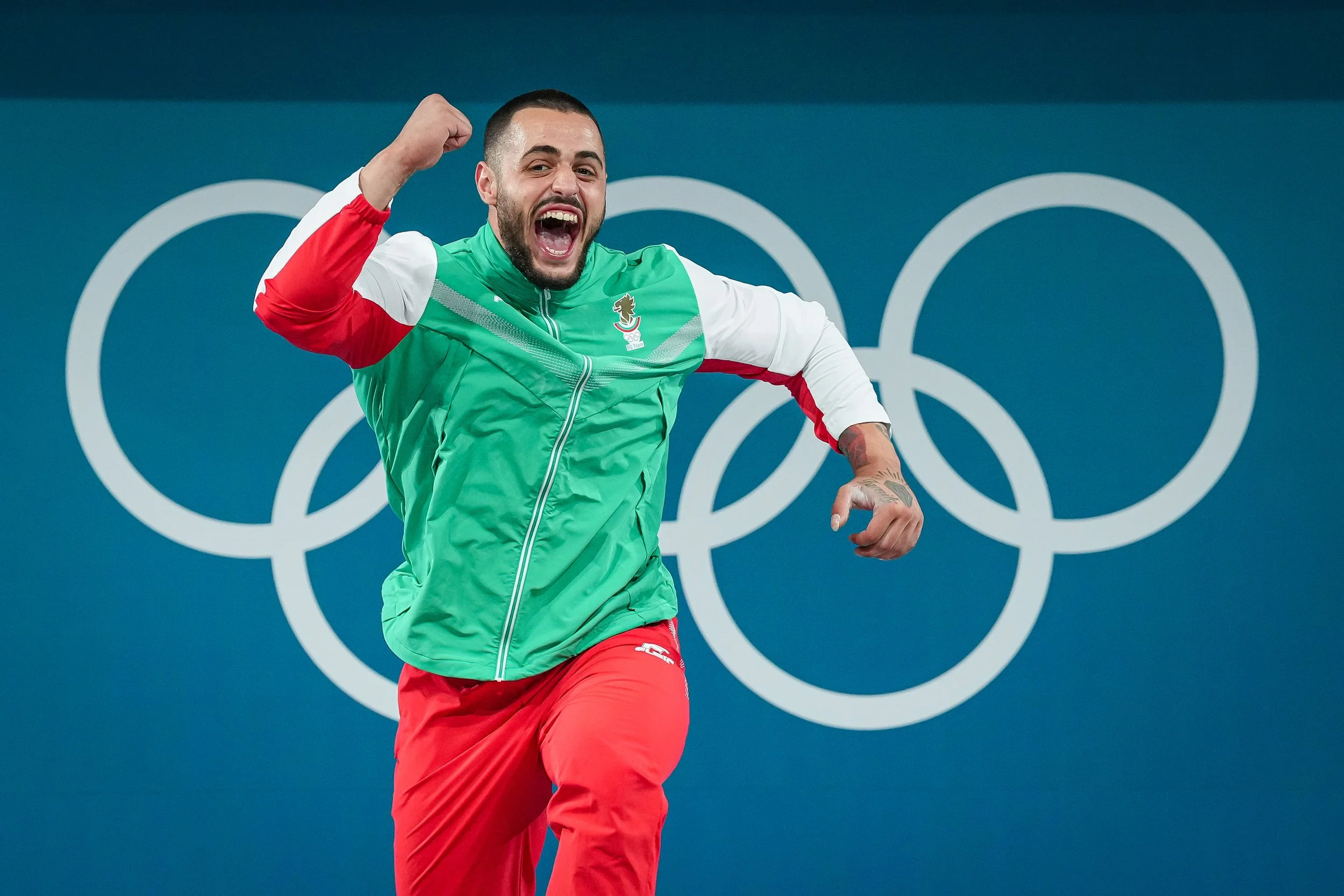 Carlos Nasar celebrating enthusiastically at the Olympics, standing in front of the Olympic rings logo.