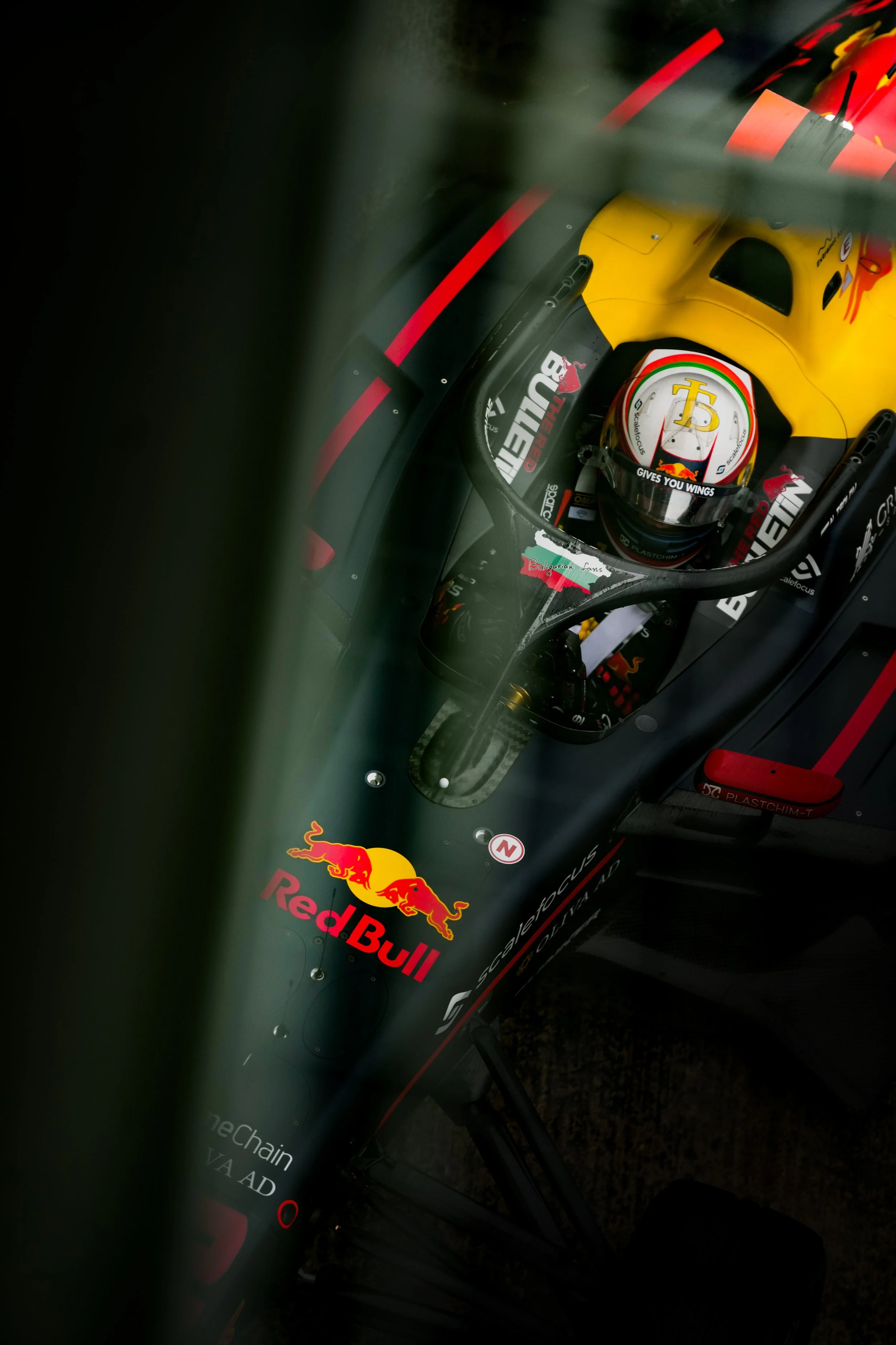 Top-down view of a racing car driver in a cockpit, wearing a helmet with red, white, and yellow colors, inside a black and yellow race car with Red Bull branding.
