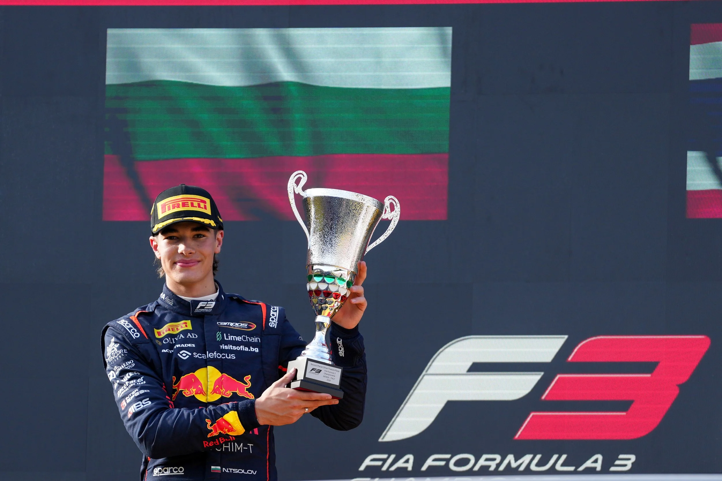 A young male F3 race car driver in a racing suit and cap, smiling and holding a trophy, standing in front of a large screen displaying the F3 logo and a Bulgarian flag.