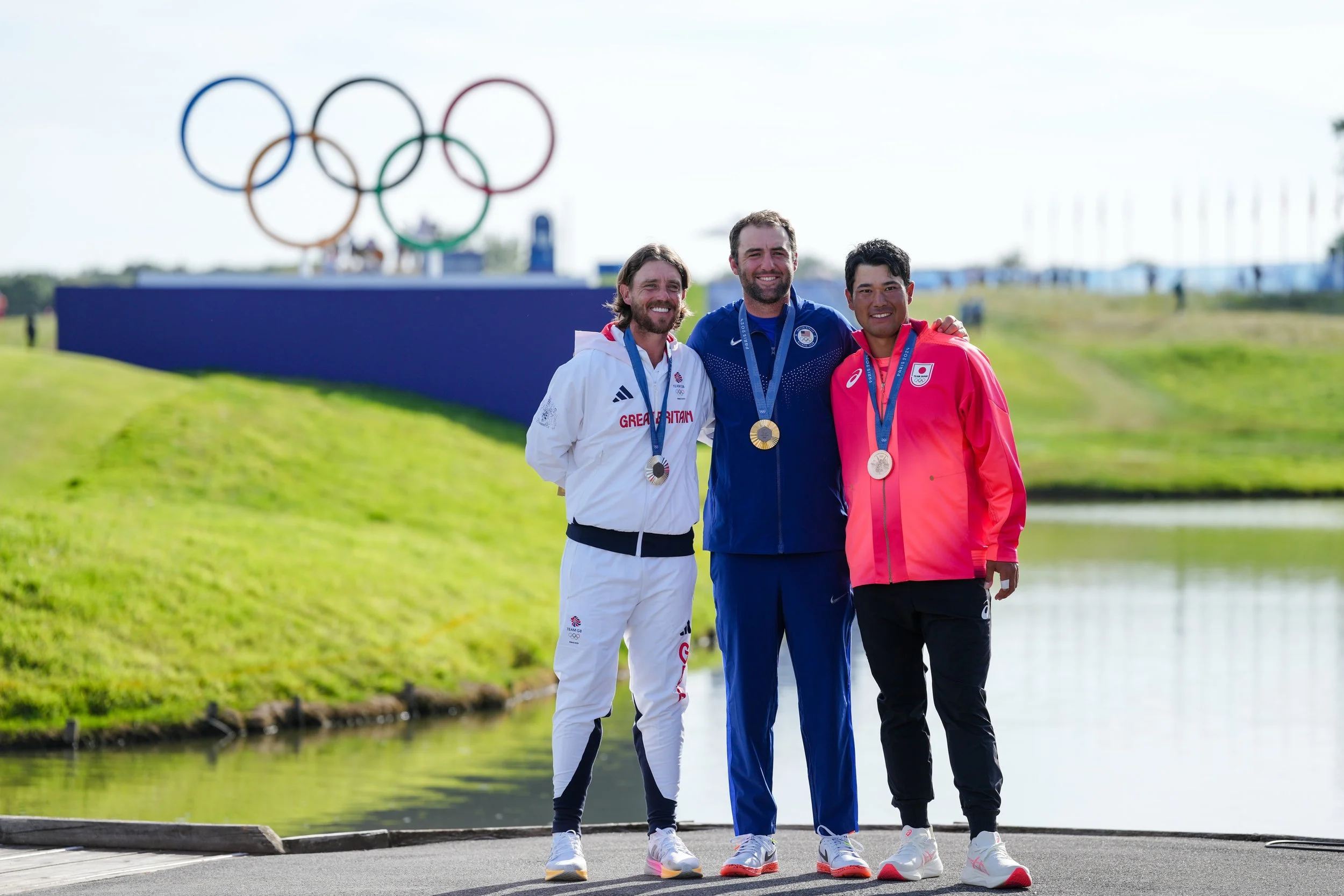 Three male athletes standing on a podium with medals, smiling, in front of the Olympic rings. They are wearing sportswear representing Great Britain, the United States, and Japan, with a body of water and grass in the background.