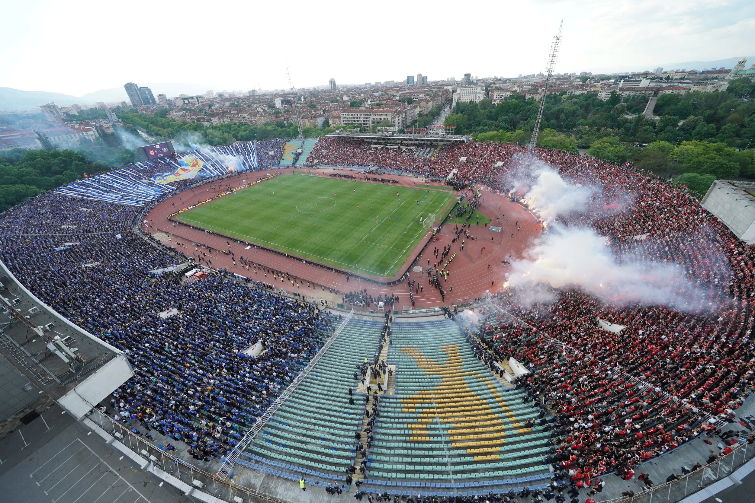 Aerial view of a soccer stadium filled with fans, some fireworks are going off, and there are stadium flags and banners.