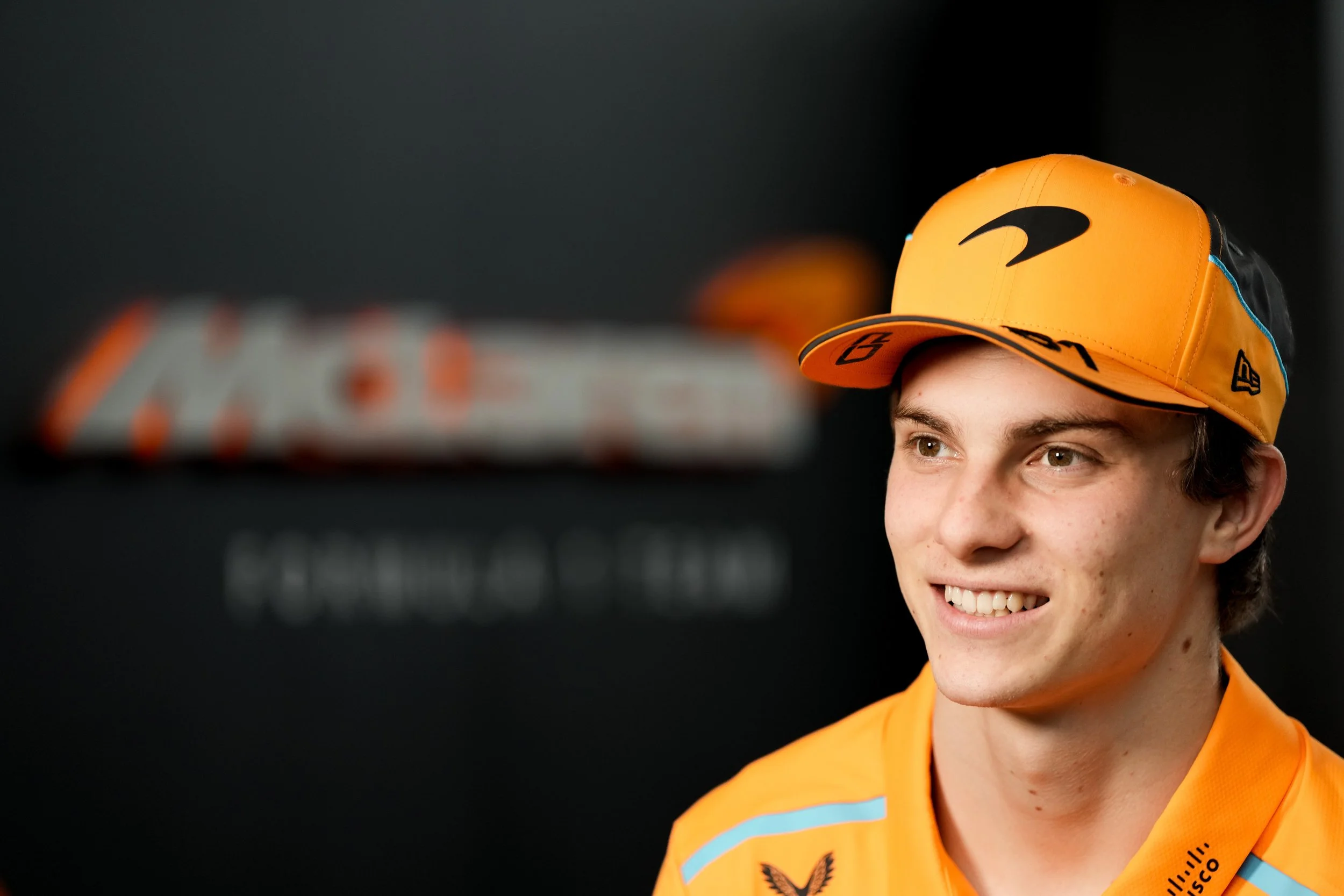 A young male race car driver in an orange cap and racing suit smiling during an interview.
