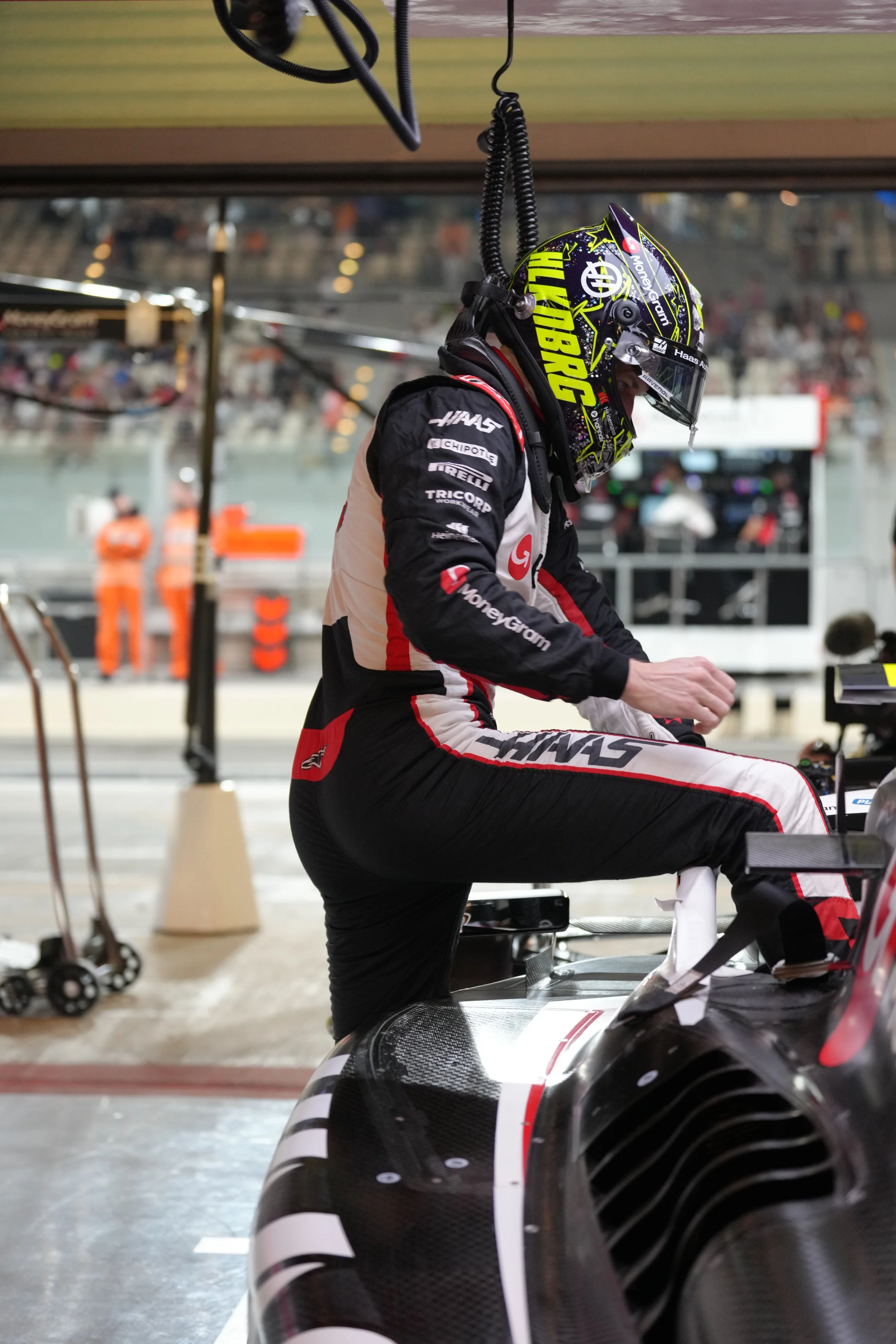 Race car driver in racing suit and helmet, preparing in the garage of a race track, with mechanics and spectators in the background.