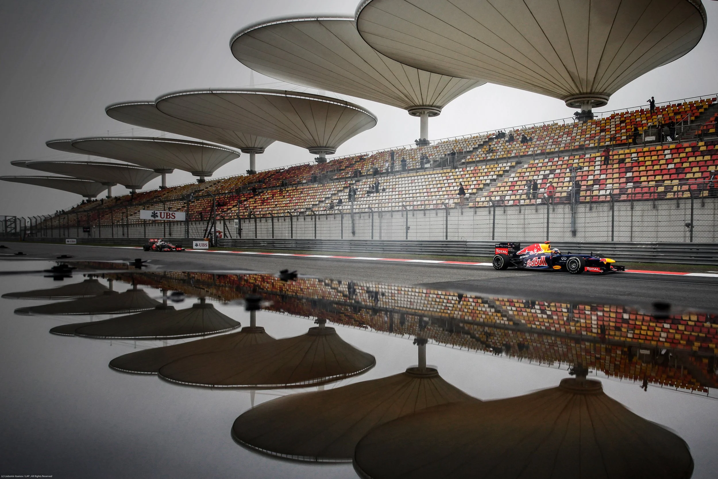 Formula 1 race car on a wet track with grandstands and unique umbrella-shaped roof structures overhead, reflected in a large puddle.