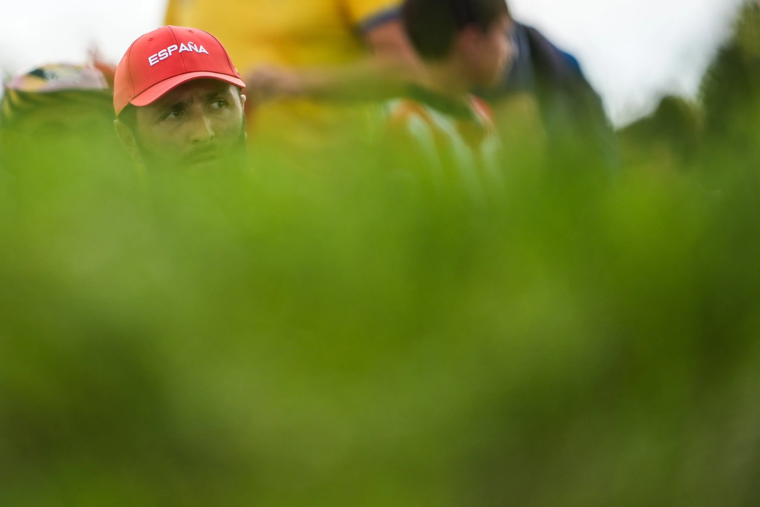 A person wearing a red cap with the word 'ESPANA' partially visible, seen through blurry green foliage, with other people in the background.
