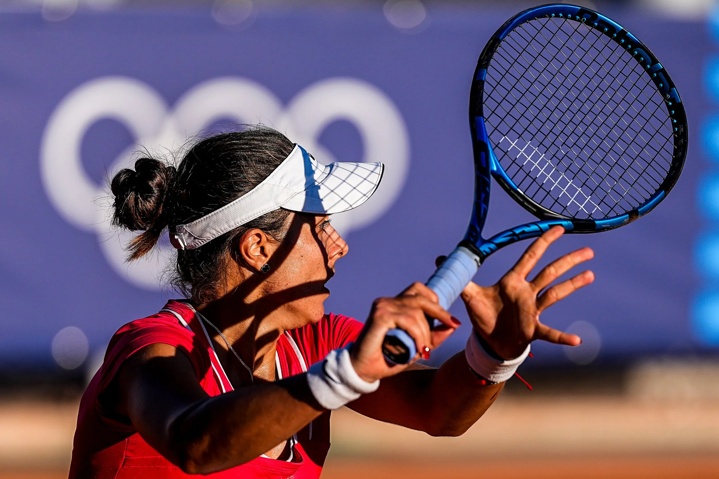 A woman tennis player wearing a red shirt and white visor is preparing to hit a tennis ball with her racket during a match.