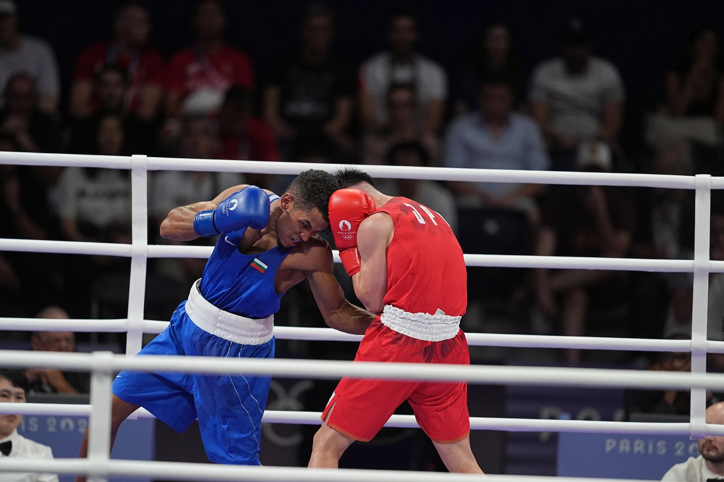 Two male boxers, one in blue and one in red, are fighting inside a boxing ring with white ropes. The boxer in blue is throwing a punch while the boxer in red is defending with his gloves up to his face.