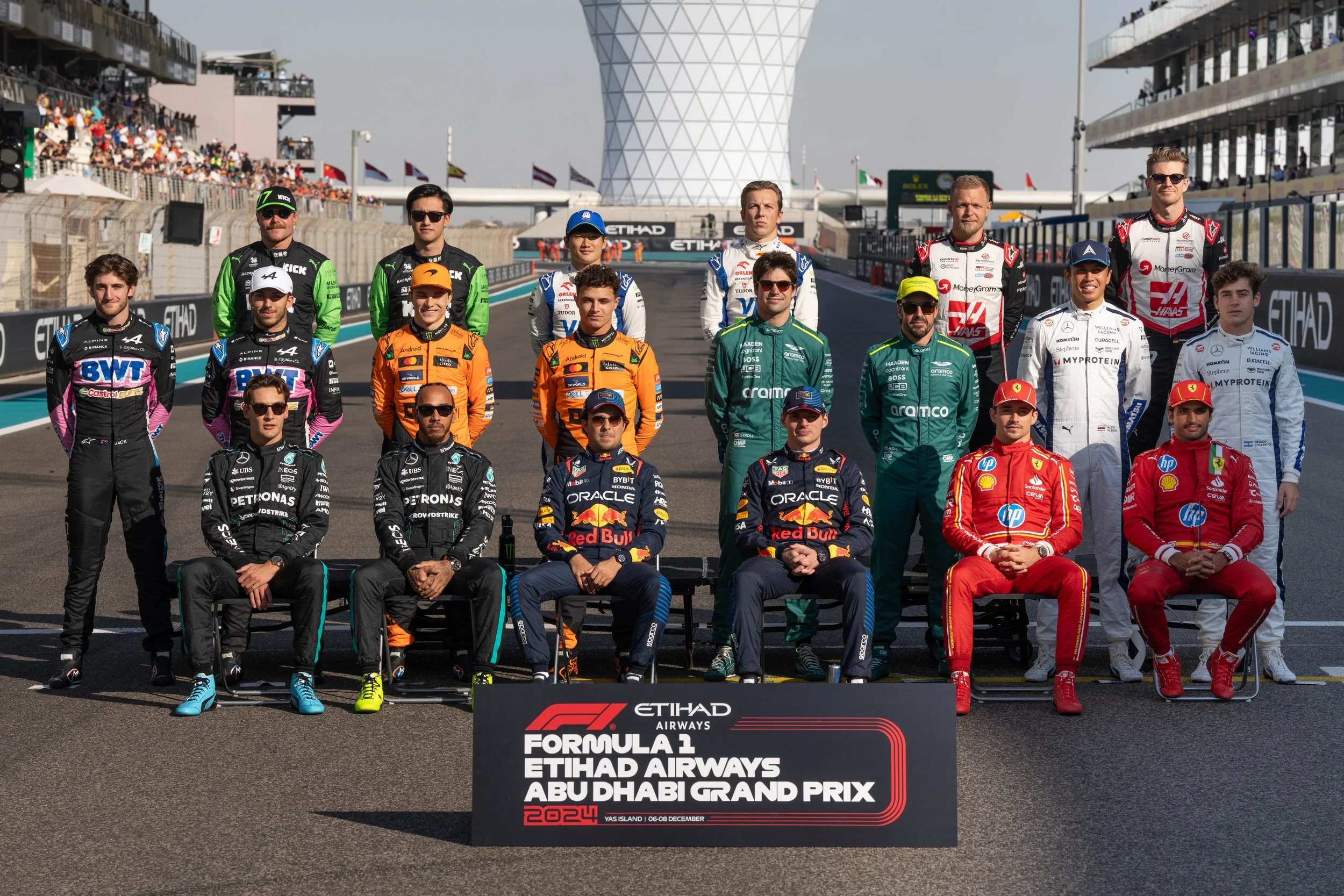 Group of Formula 1 drivers and team members posing on the starting grid at the Abu Dhabi Grand Prix, held at Yas Island in December 2024, with a large sponsor banner at the front of the image.