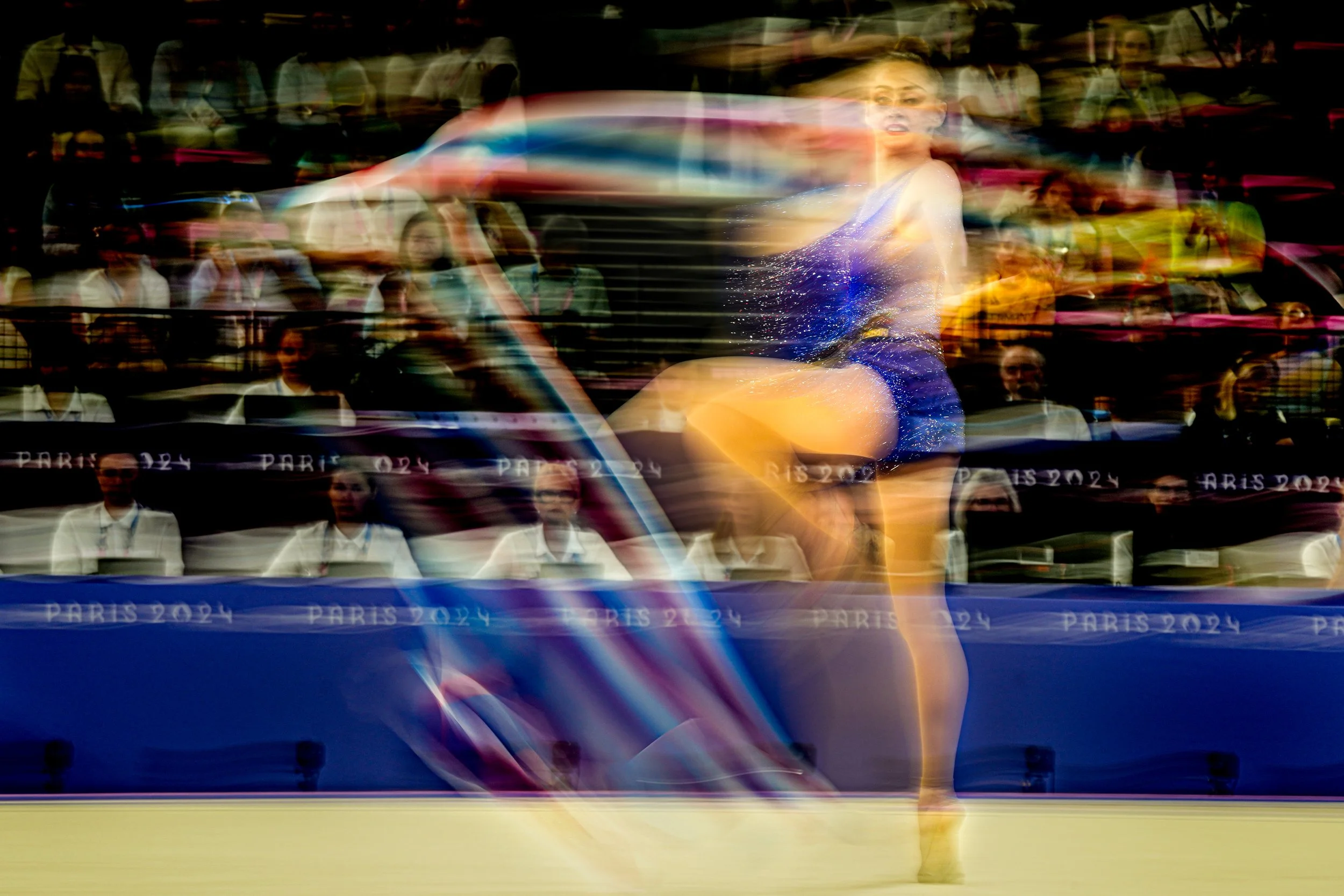 A rhythmic gymnast performing a dance routine in a blue costume on a stage at Paris 2024 event, with motion blur showing her spinning and leaping, and an audience seated behind her.