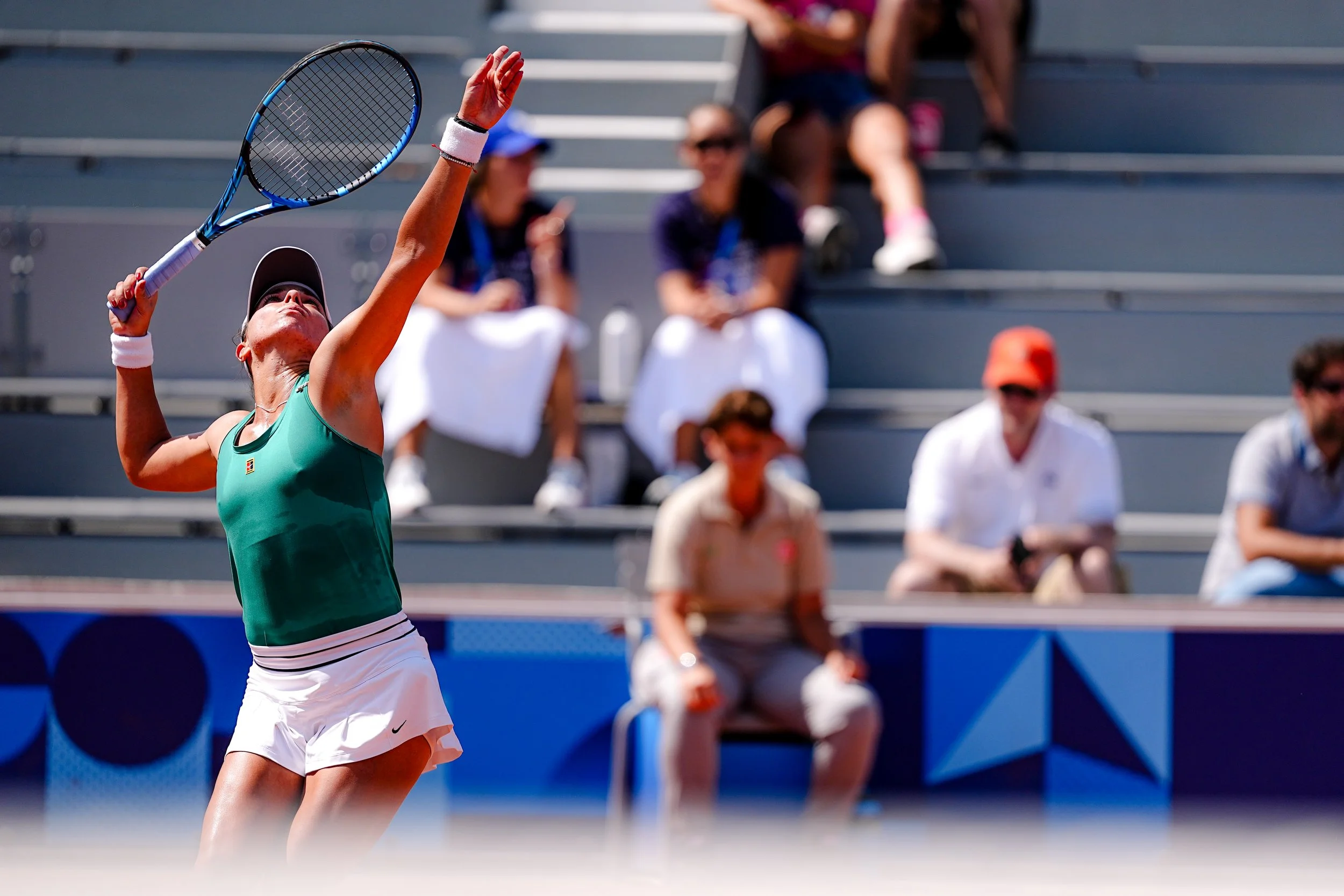A female tennis player in a green top and white skirt preparing to serve on a tennis court, with spectators sitting on bleachers in the background.