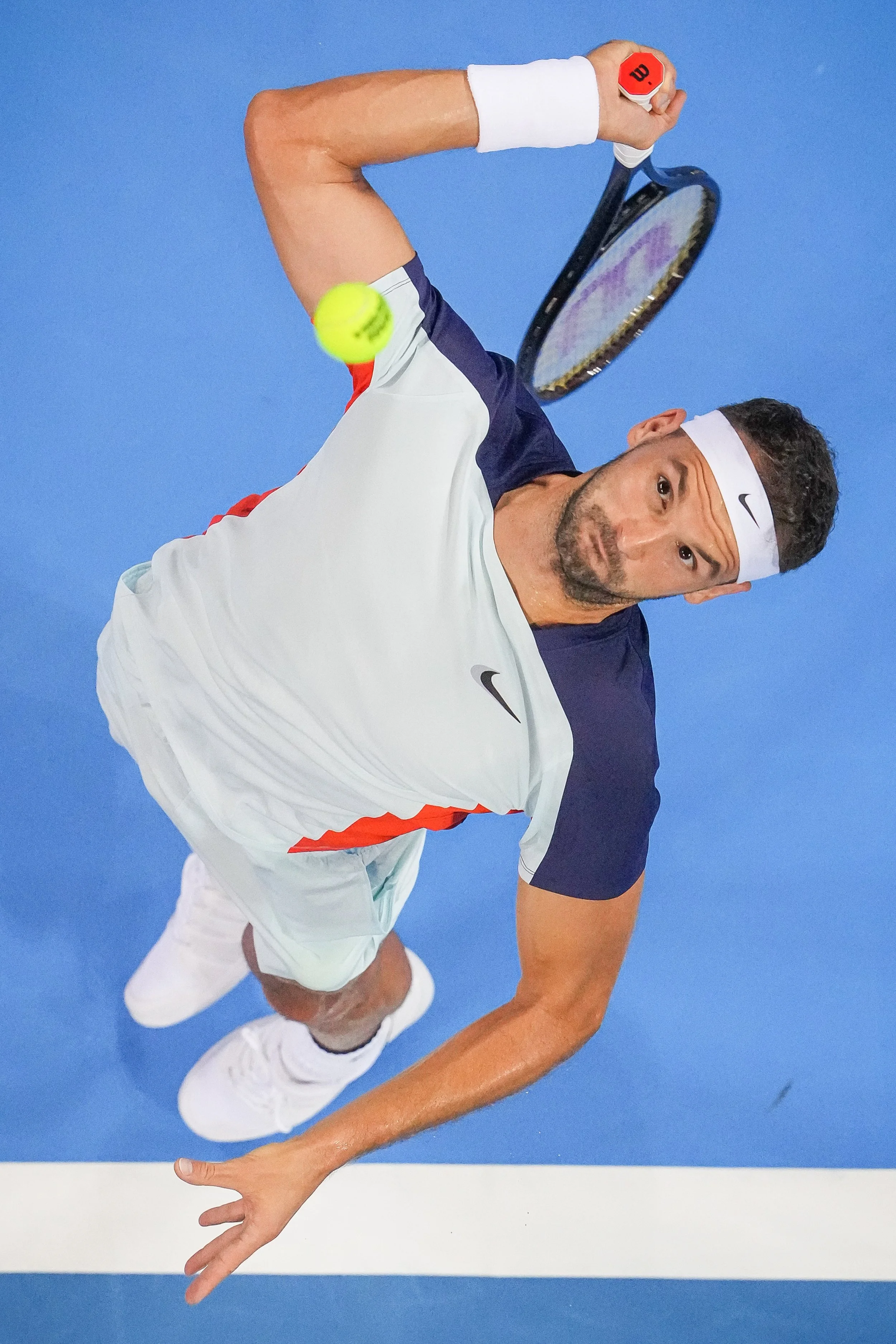 A male tennis player wearing a white and navy sports outfit, white headband, and wristband, preparing to hit a tennis ball with his racket on a blue tennis court.