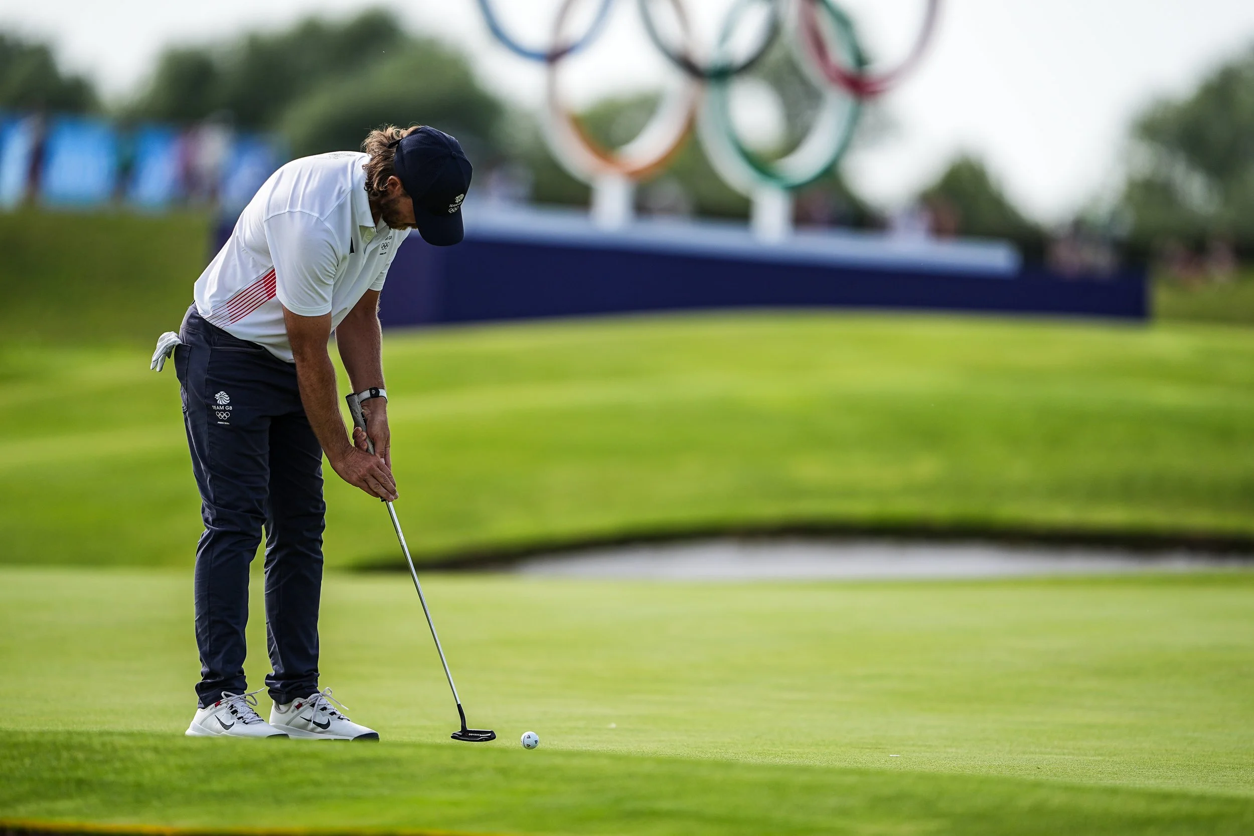 A golfer prepares to putt on a golf course with the Olympic rings in the background during the Olympics.