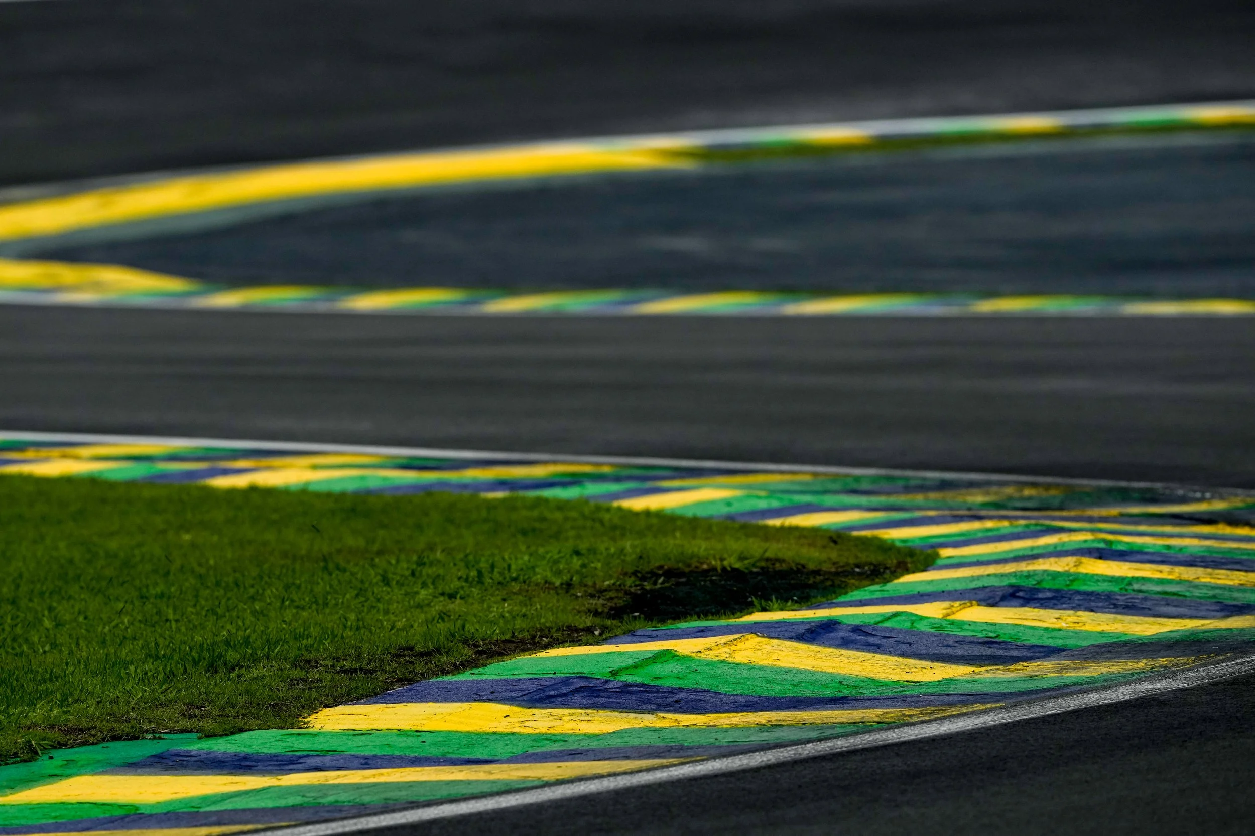 A section of a race track with green, yellow, and blue painted curbs, and a grassy area near the edge of the pavement.