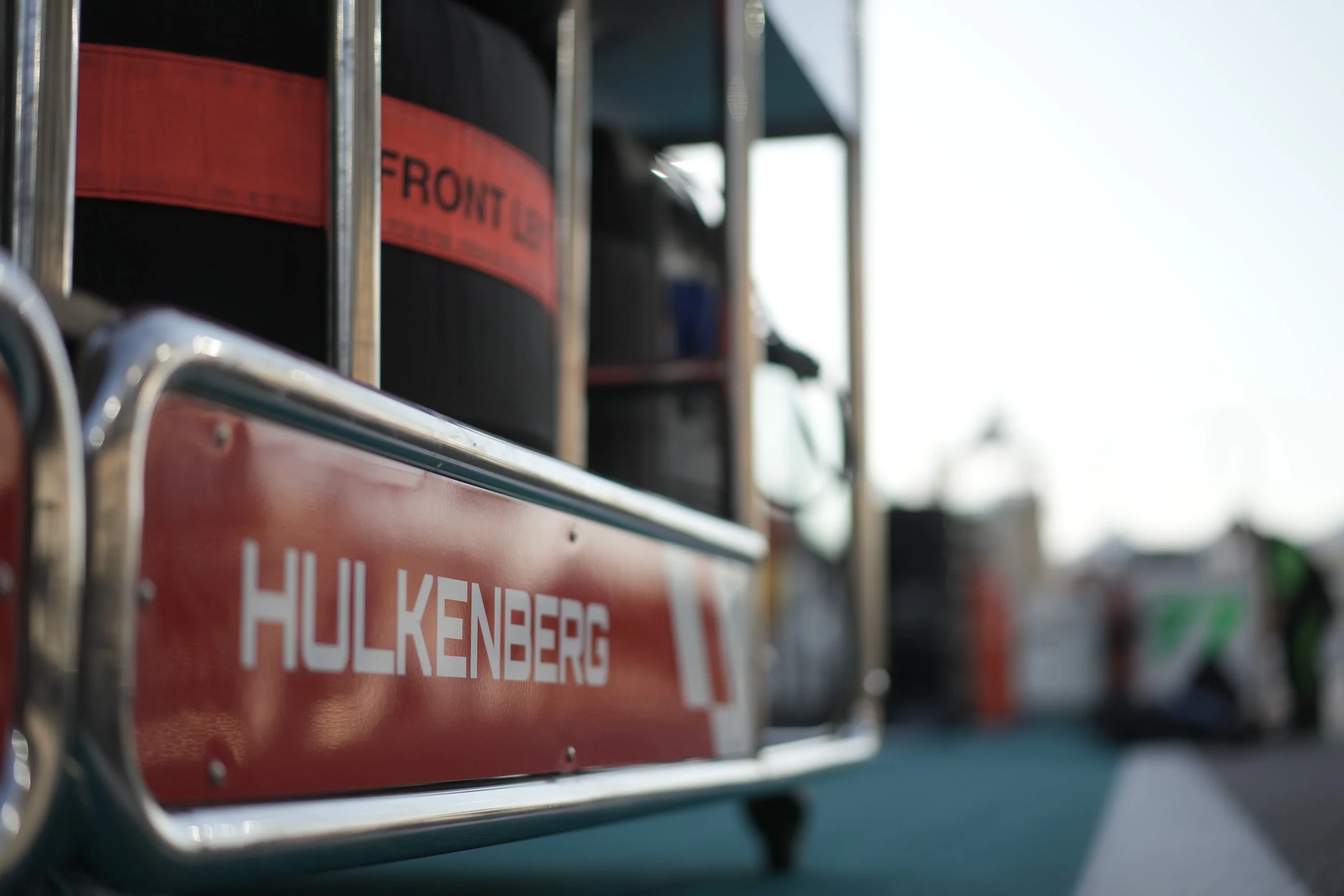 Close-up of a red and silver Hiltonberg branded sign on a metal cart, with shelves holding black containers and other equipment in the background on a outdoor street or parking lot.
