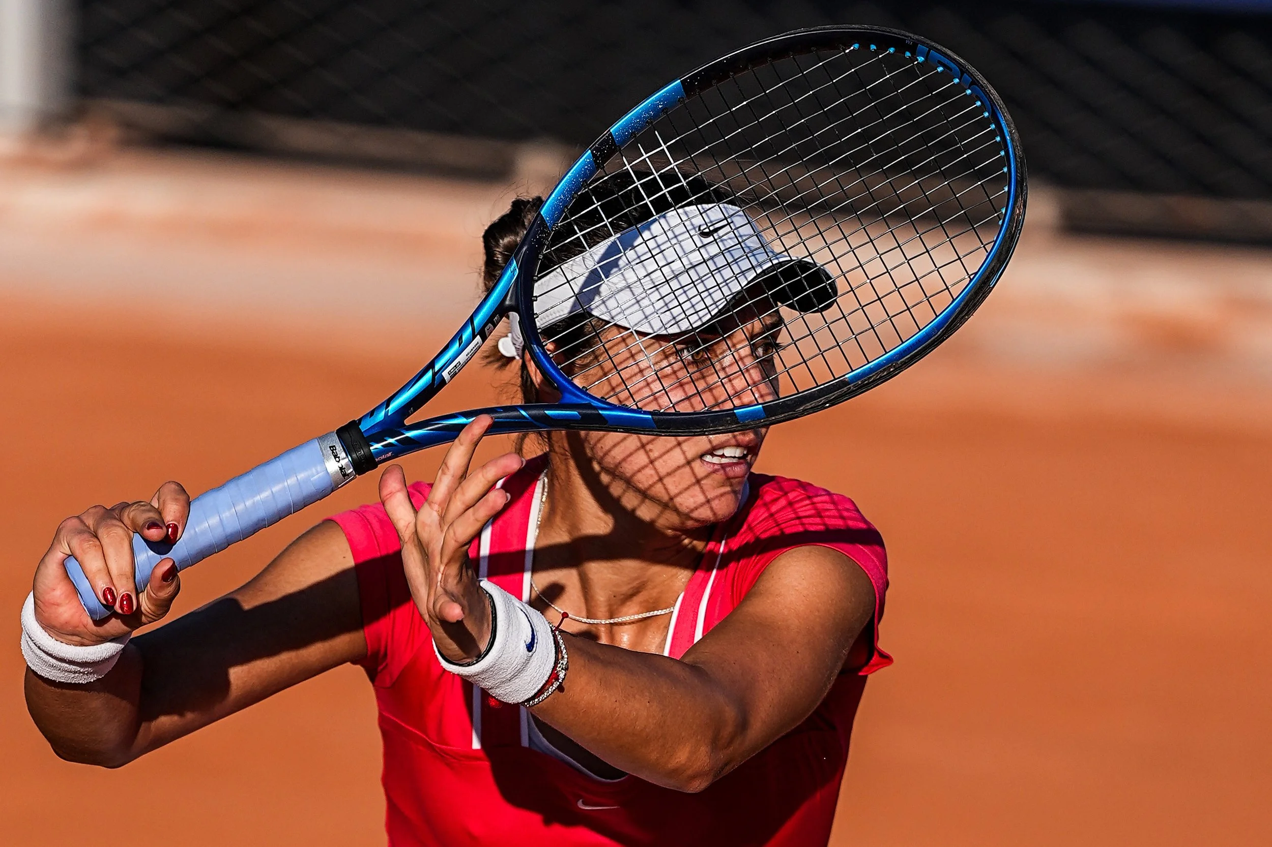 Tennis player with a red shirt and white cap holding a tennis racquet on a clay court.