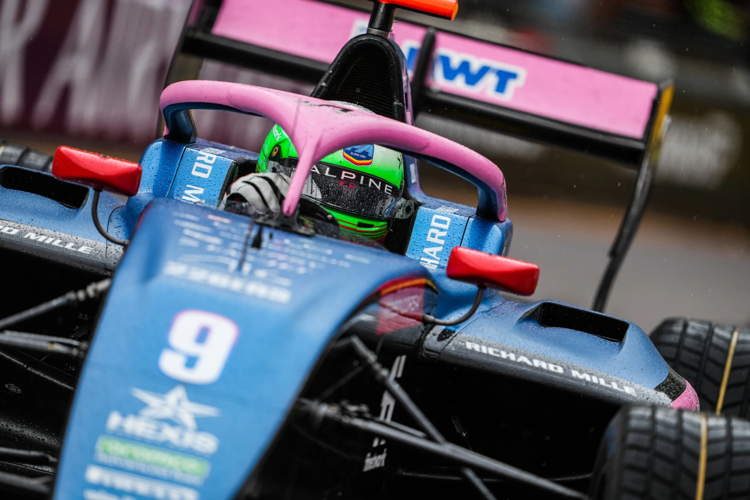 Close-up of a blue race car with the number 9 on the front, rain droplets on its surface, and a driver in a green helmet sitting inside. The car features a pink roll hoop and red side mirrors.