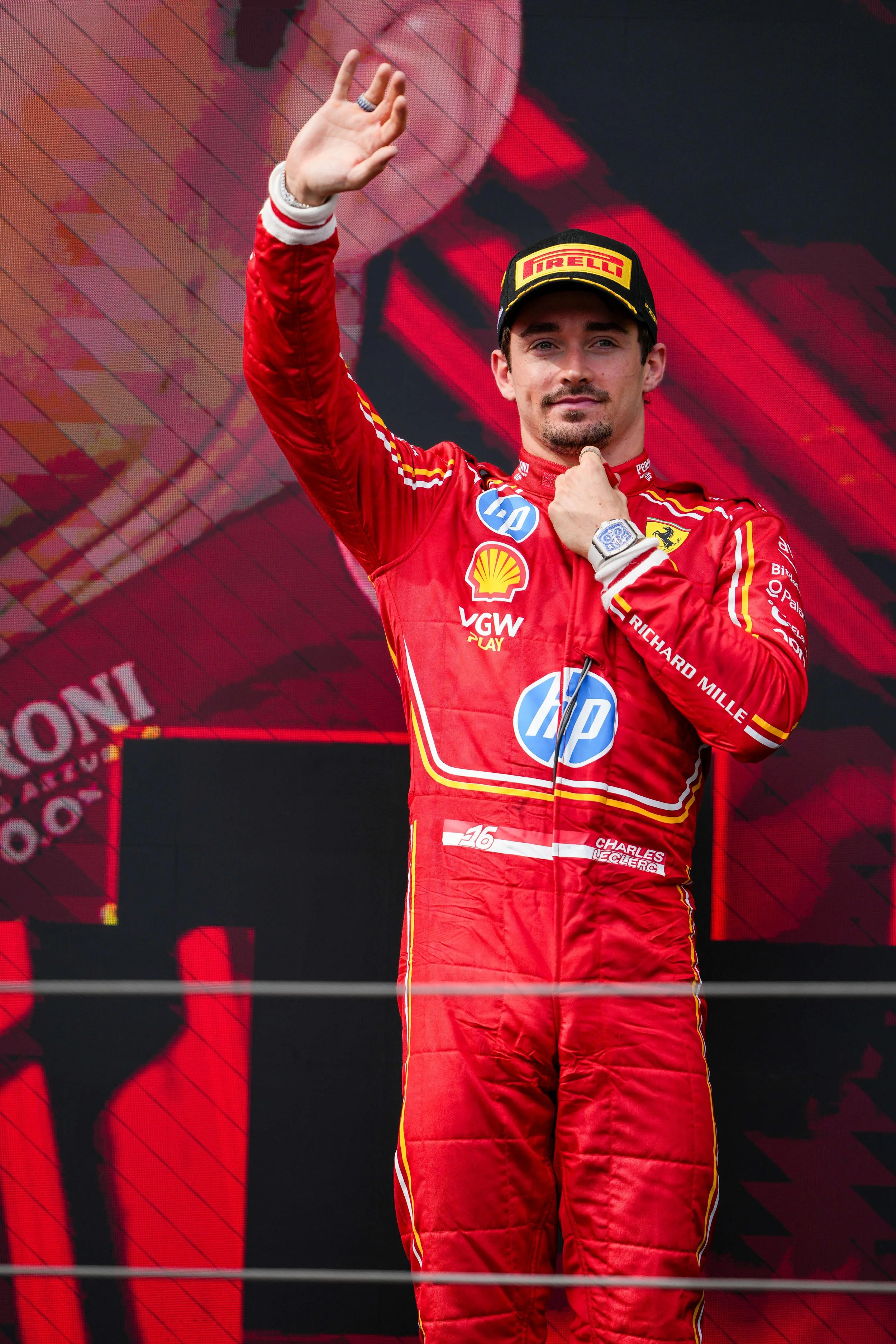 Race car driver in red racing suit waving to the crowd on podium, with sponsor logos and cap, celebrating a race win.