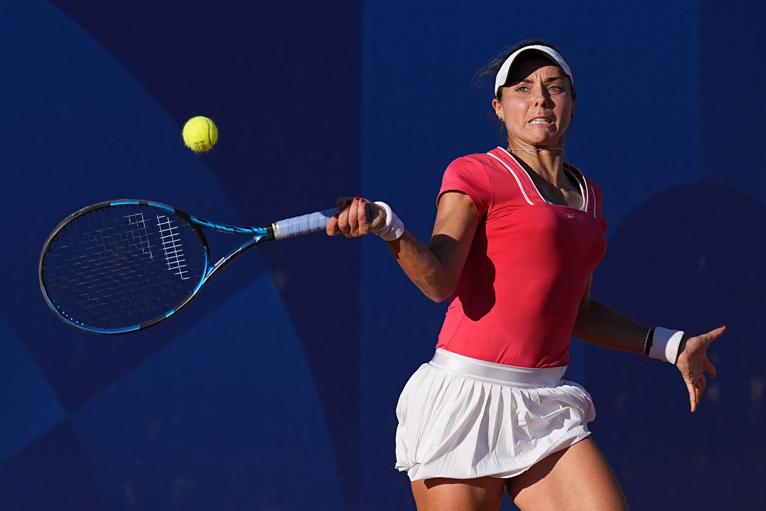 Victorina Tomova in red shirt and white skirt hitting a tennis ball with a racket.